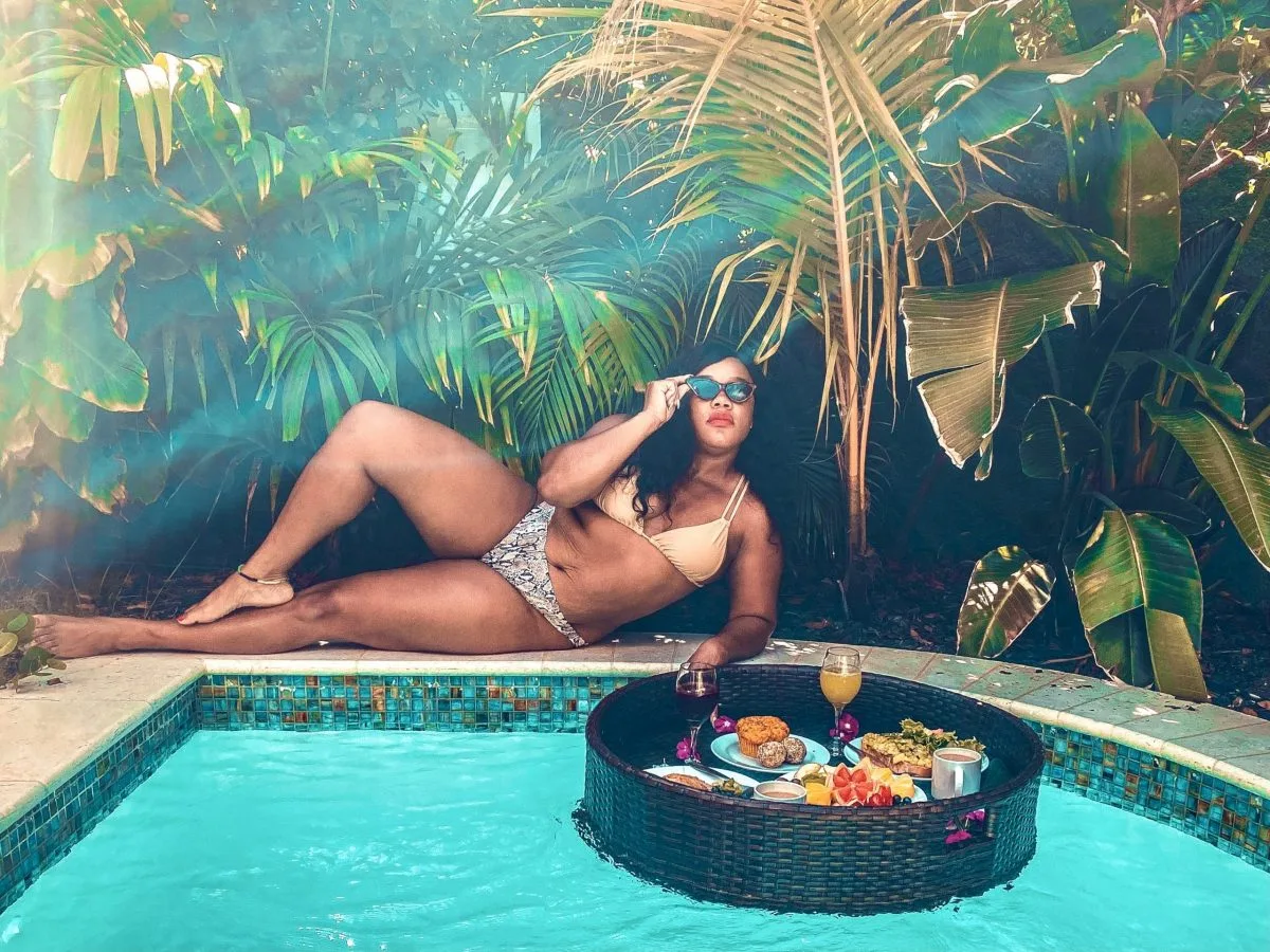 Marquita lounges poolside in a bikini at Boardwalk Boutique Hotel in Aruba, with a floating breakfast tray in front of her featuring tropical fruits, pastries, and drinks. Lush palm leaves frame the scene, creating a serene, upscale vibe that captures where to stay in Aruba for a private, boutique experience.