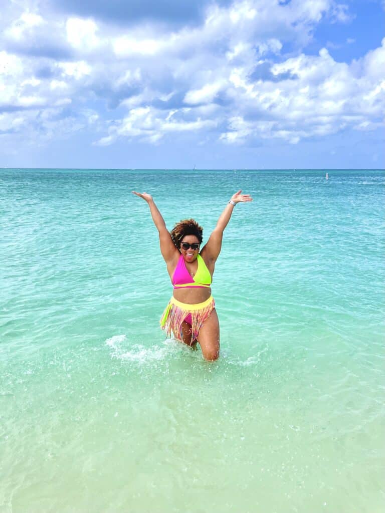 Splashing water in Eagle Beach, Aruba wearing a bright pink and green 2 piece swimsuit with a multi-color coverup skirt