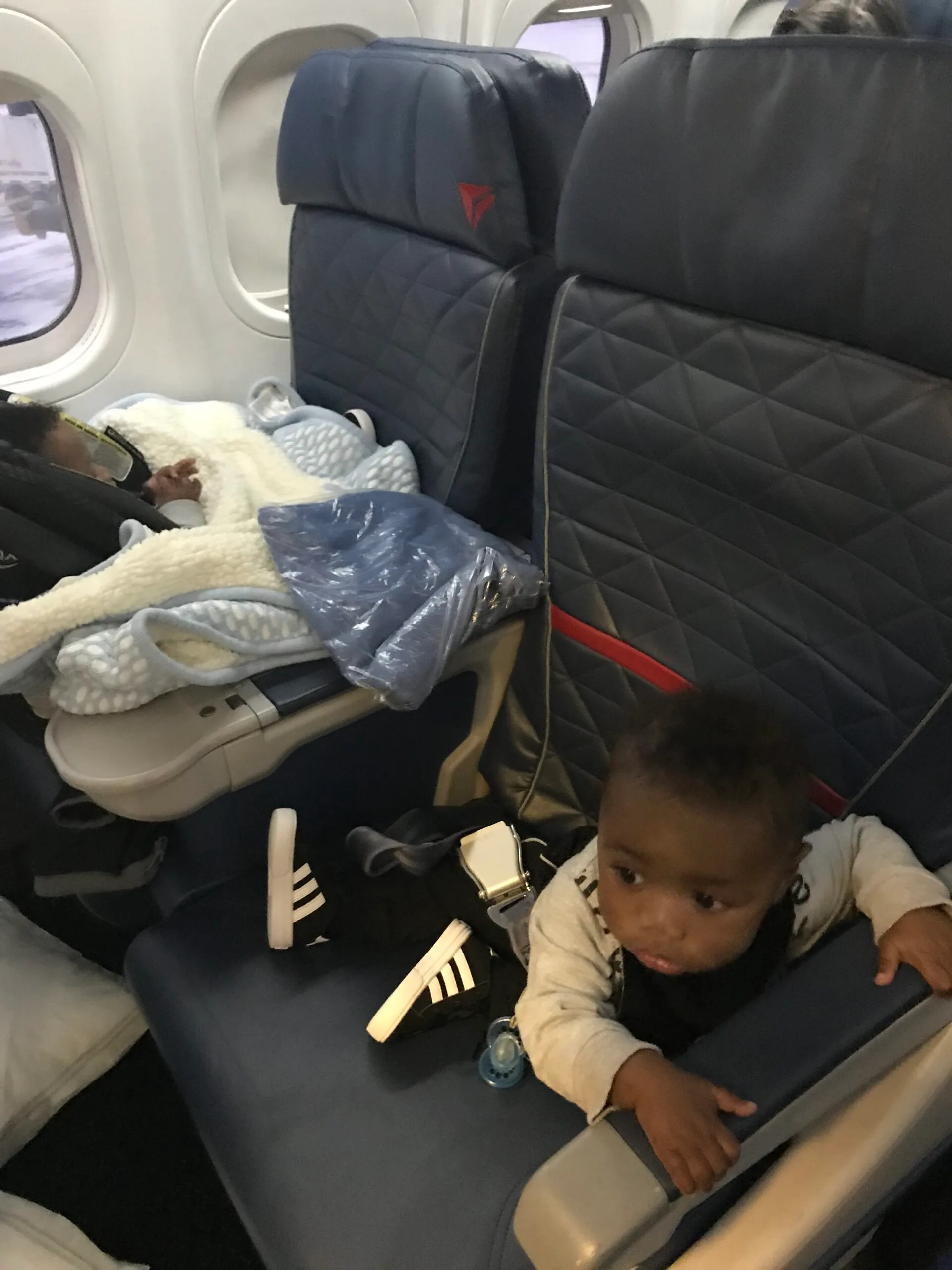 Twin babies sitting on the plane- one in a car seat while the other is sitting in a seat.