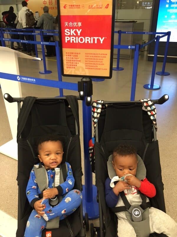 Twin boys in a travel stroller in front of a Sky Priority sign at a Chinese airport