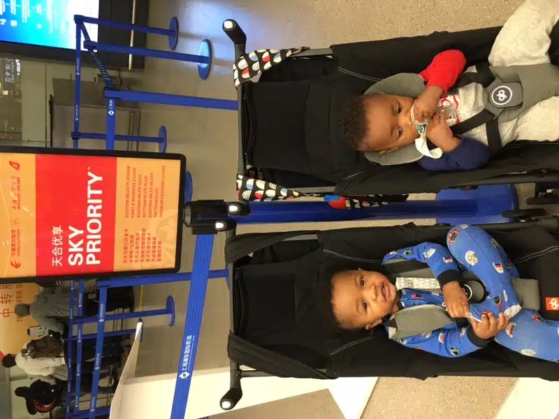 Twin boys in a travel stroller in front of a Sky Priority sign at a Chinese airport