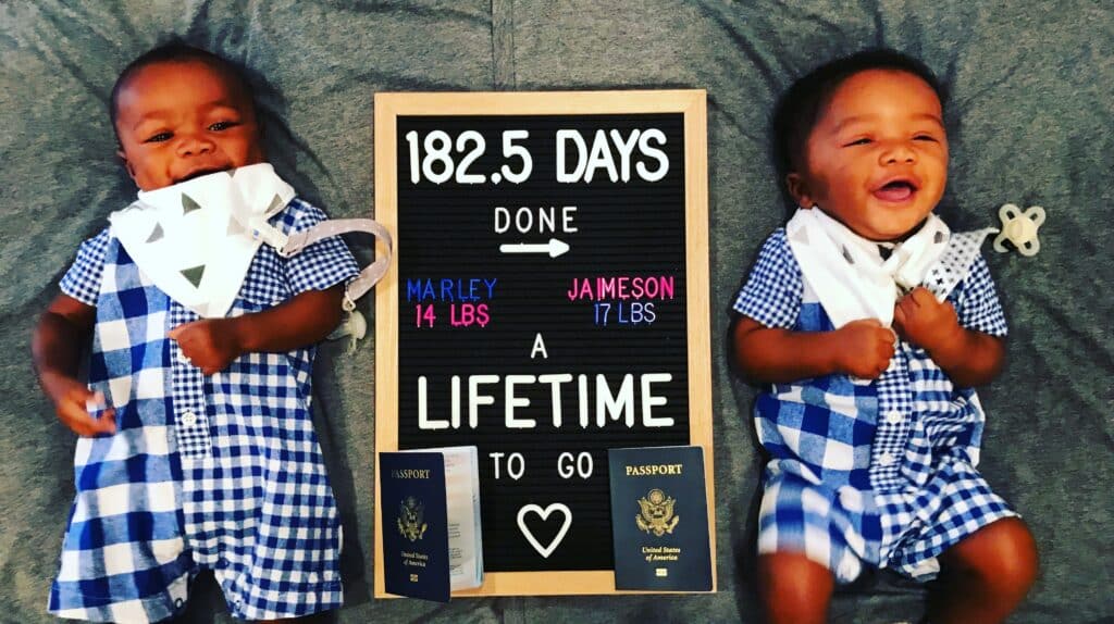 Twin baby boys laying on a grey blanket with a 6 month birthday sign and their passports