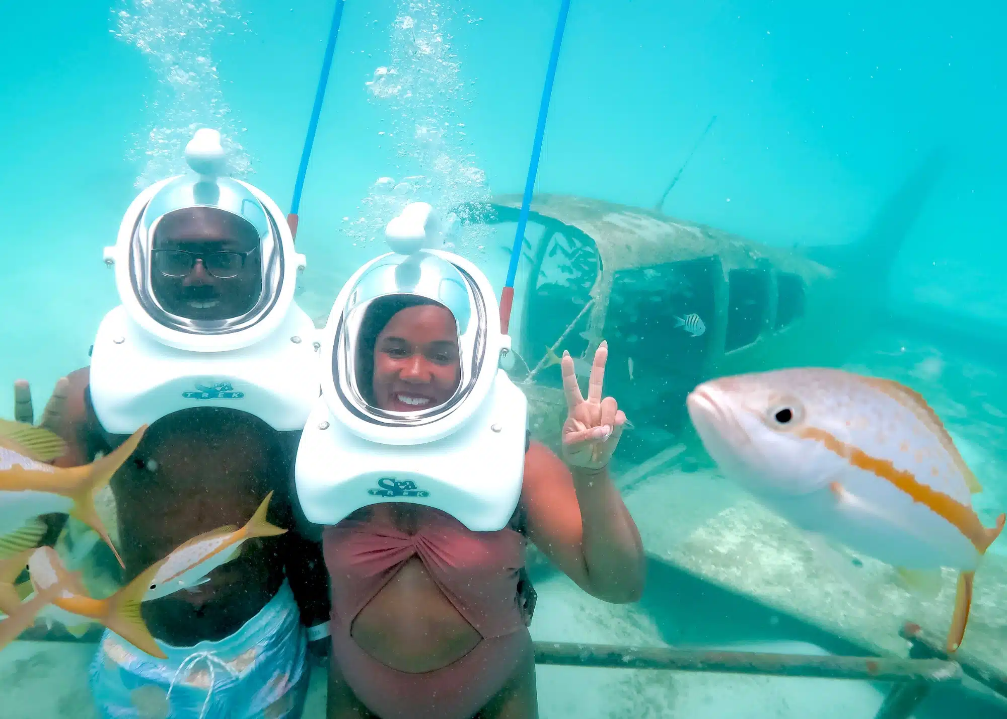 Marquita and her husband explore the ocean floor during the Sea Trek underwater helmet walk in Aruba, posing in front of a sunken airplane surrounded by tropical fish. Both wear clear dome helmets as they enjoy the unique underwater experience.