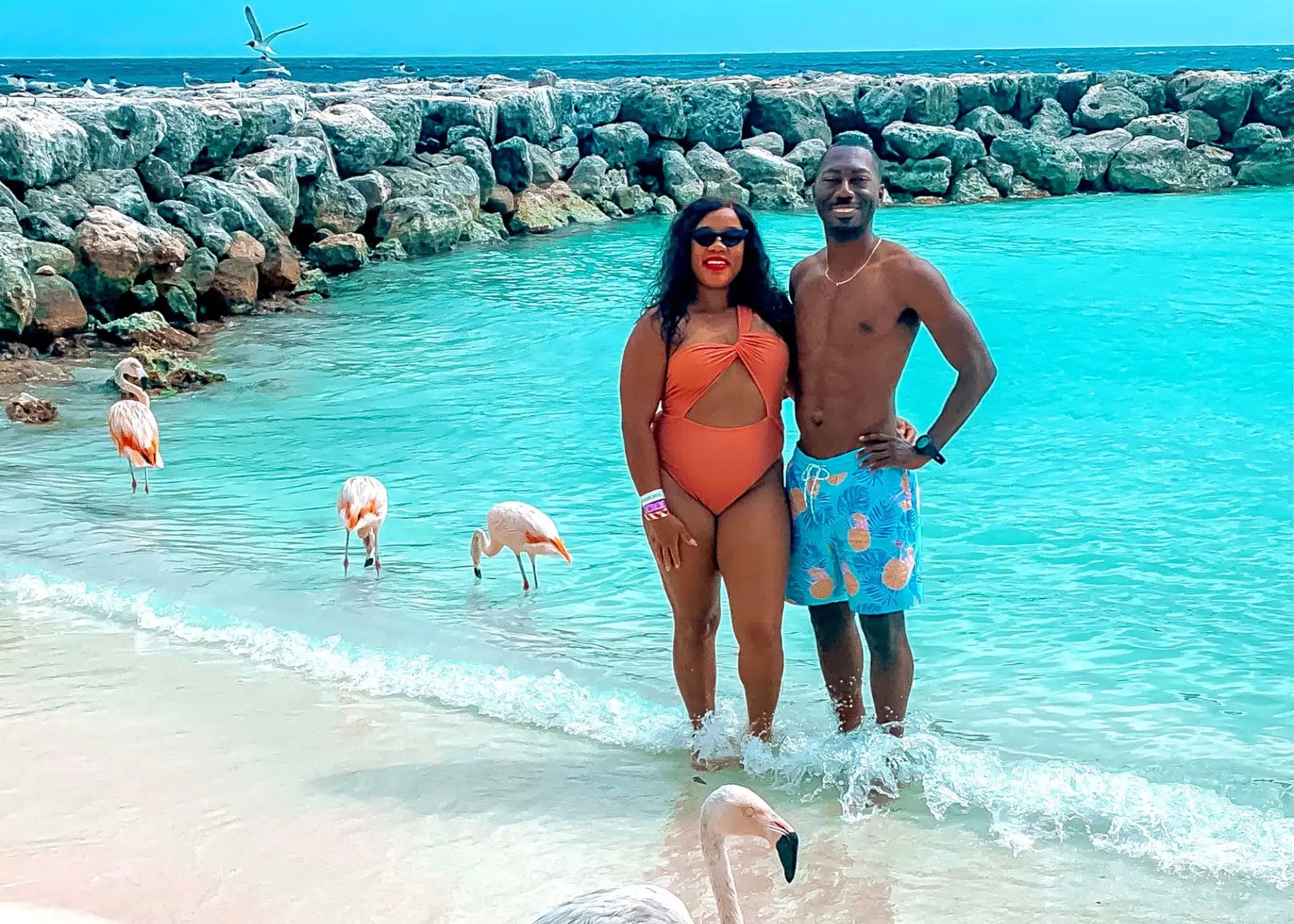 Marquita and her husband pose at the edge of the crystal-clear water on Flamingo Beach in Aruba, surrounded by a group of flamingos wading along the shore. Behind them, a rocky barrier and turquoise sea set the scene for a perfect tropical moment.