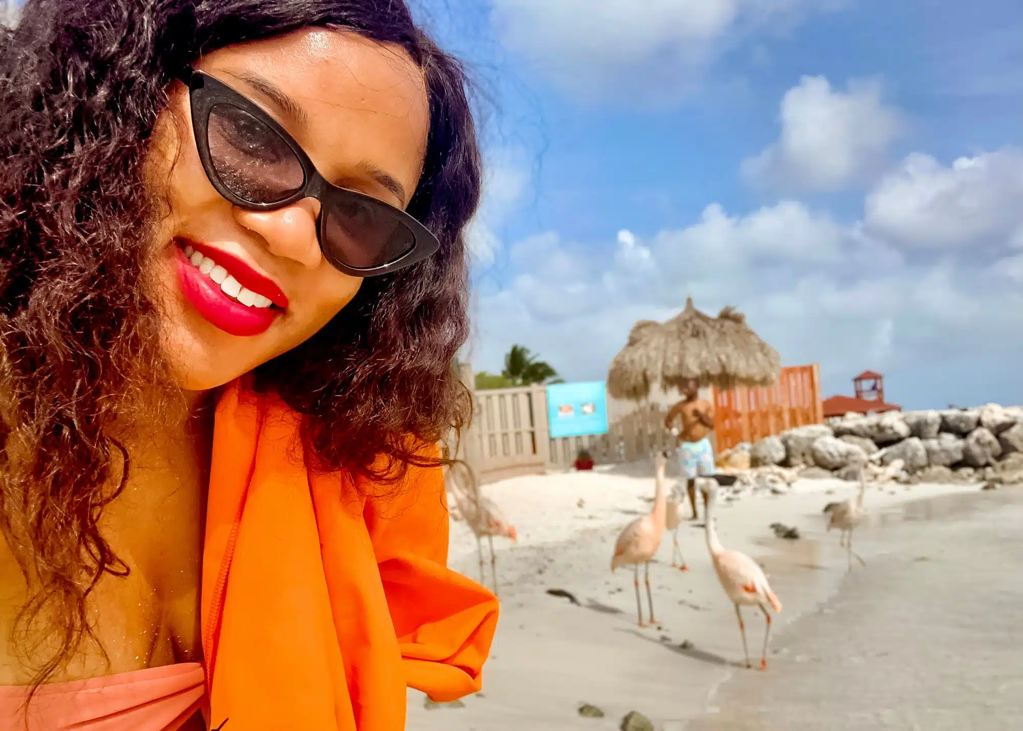 Marquita takes a bright, cheerful selfie at Flamingo Beach in Aruba, wearing cat-eye sunglasses and a coral swimsuit with an orange cover-up. Behind her, flamingos walk along the shoreline near thatched umbrellas and colorful cabanas under a partly cloudy sky.