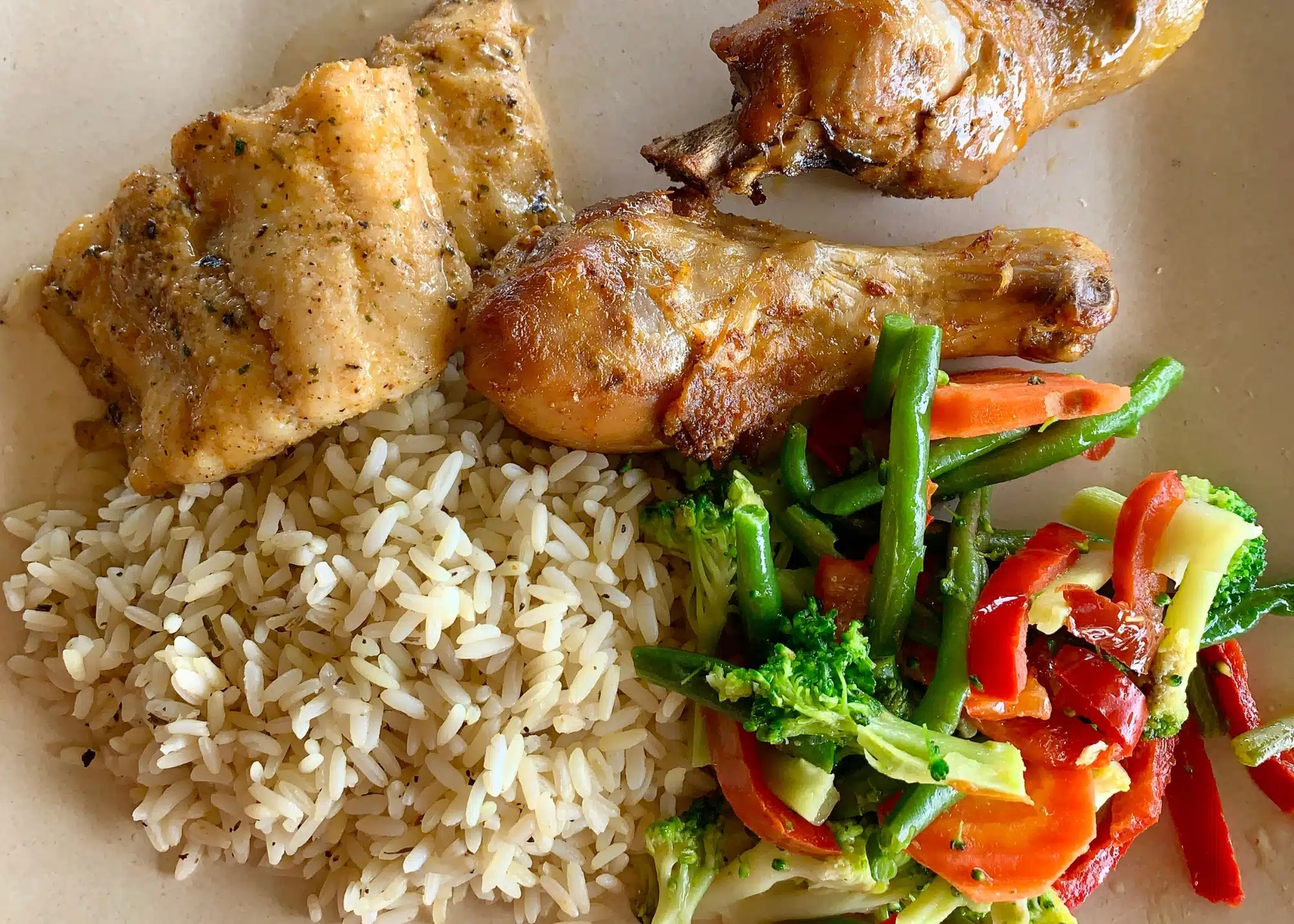 A plate of food included with the De Palm Day Pass in Aruba featuring seasoned grilled chicken drumsticks, a fillet of lightly spiced fish, a scoop of herbed rice, and a colorful medley of sautĂŠed vegetables including broccoli, carrots, red peppers, and green beans.
