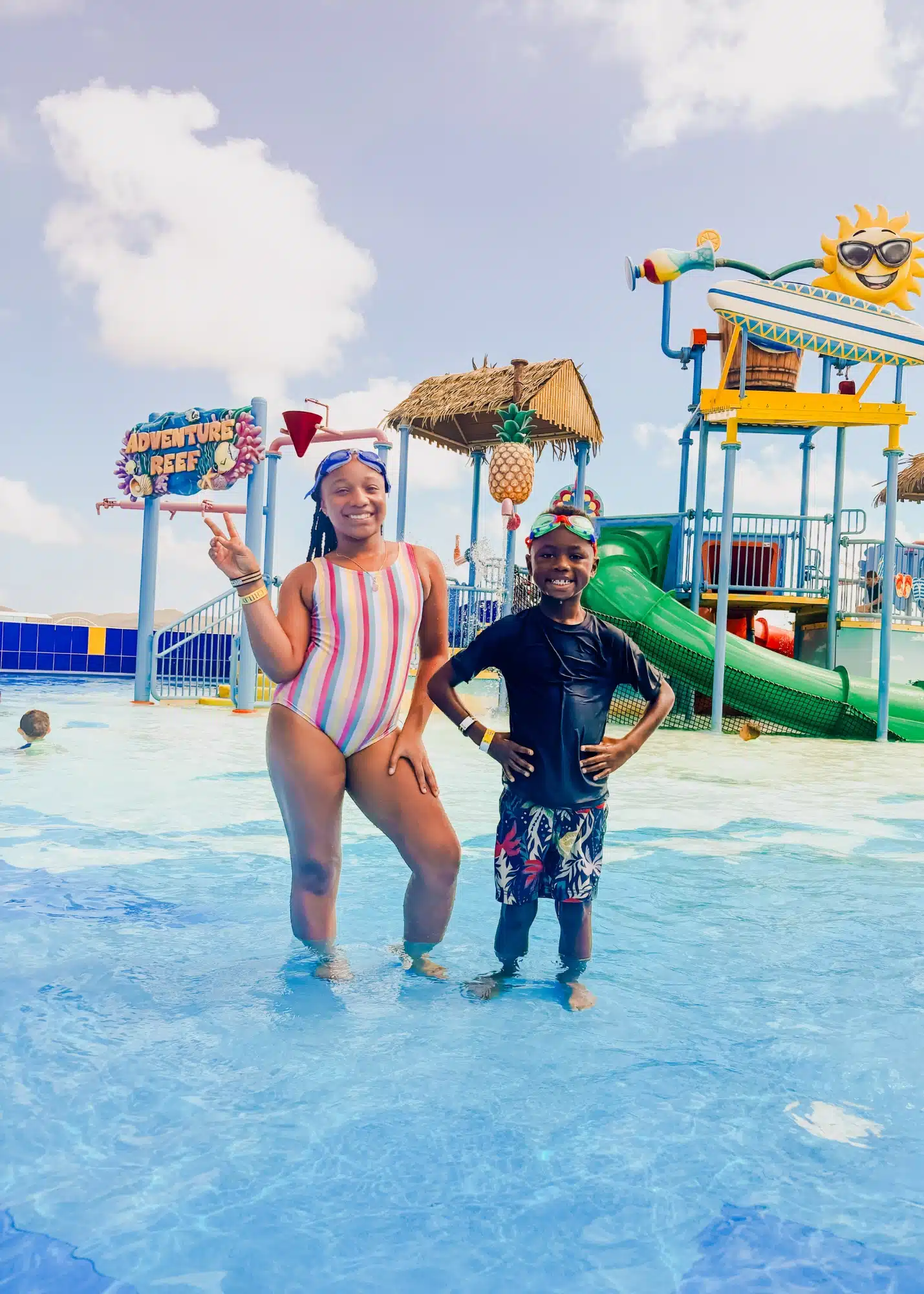 Two kids smile and pose in the shallow splash area at Adventure Reef on De Palm Island, Aruba. Behind them, colorful water slides, playful tropical-themed decor, and a giant sunglasses-wearing sun add to the fun, family-friendly atmosphere.