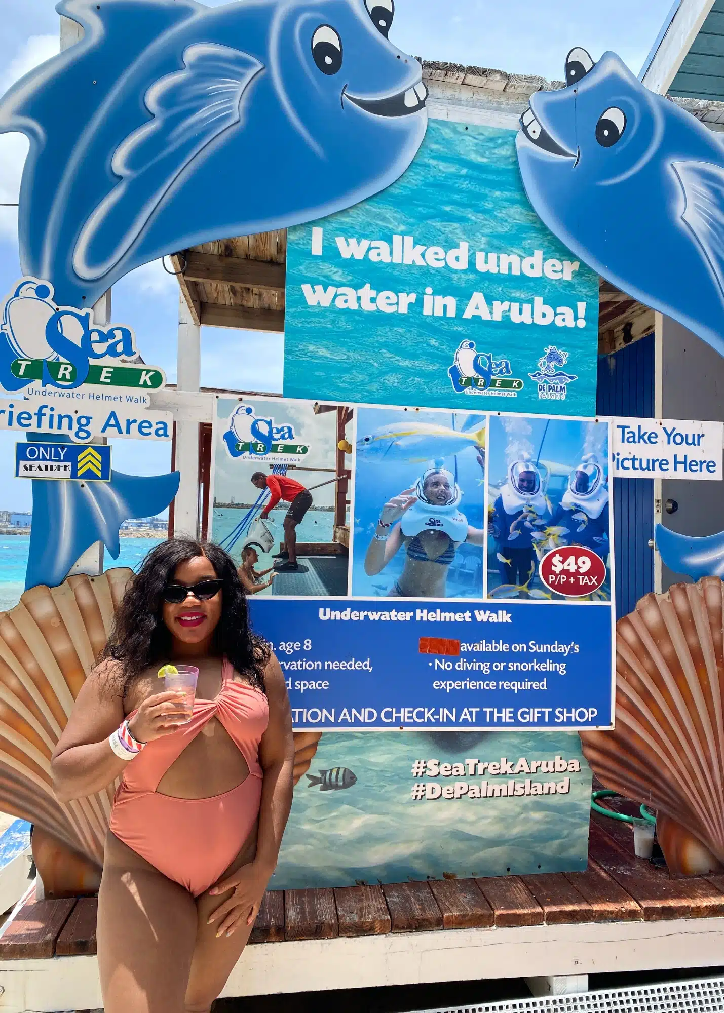 Marquita stands in front of the Sea Trek Aruba sign at De Palm Island, holding a drink and wearing a peach-colored swimsuit. The colorful sign behind her promotes the Underwater Helmet Walk experience with the phrase âI walked under water in Arubaâ and photos of participants exploring underwater with helmets.