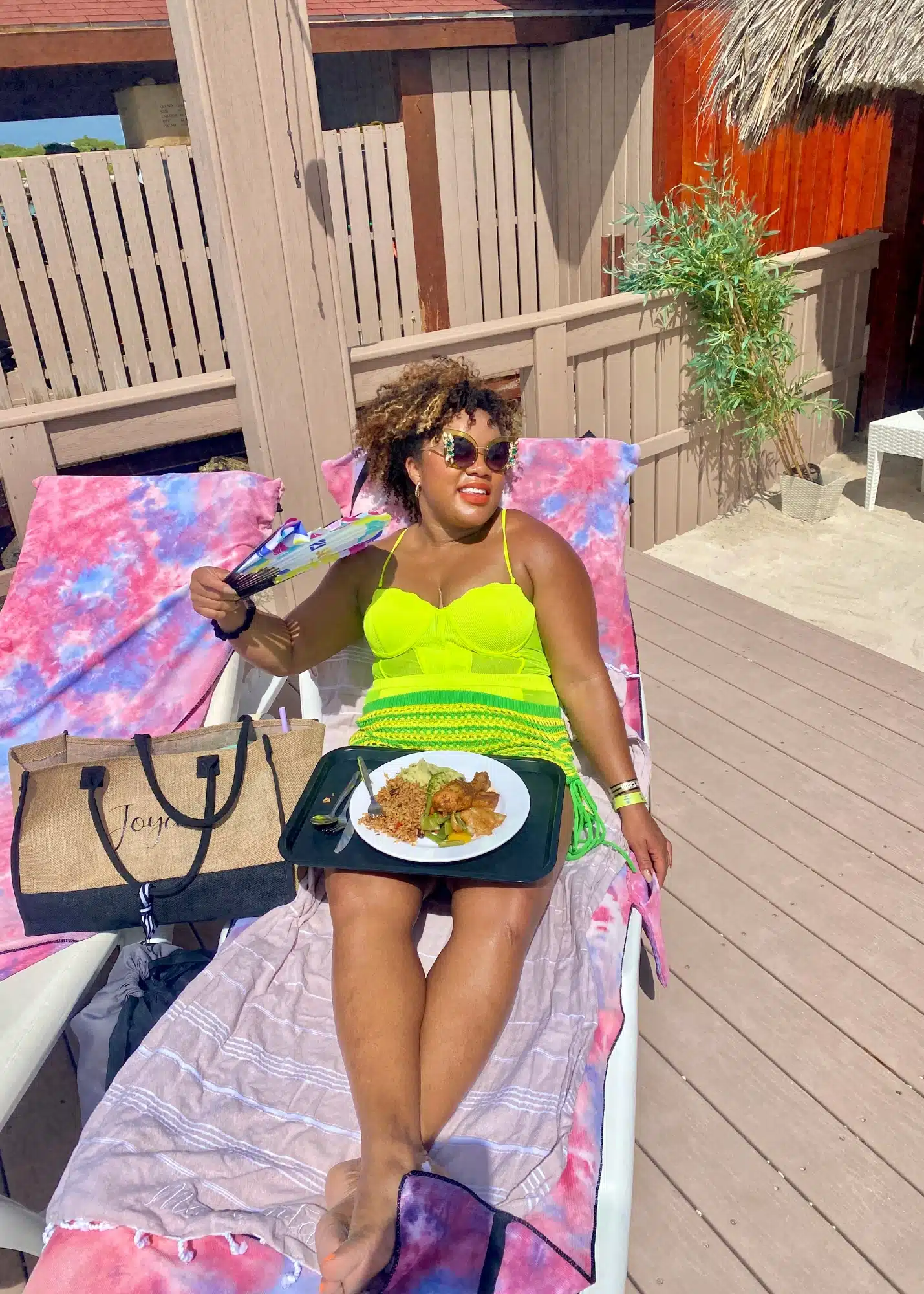 Marquita lounges on a beach chair at Flamingo Beach in Aruba, wearing a bright neon green swimsuit cover-up and colorful sunglasses while fanning herself with a floral hand fan. A plate of food from the De Palm Island day pass rests on her lap as she relaxes under the sun on a pink and purple tie-dye towel.