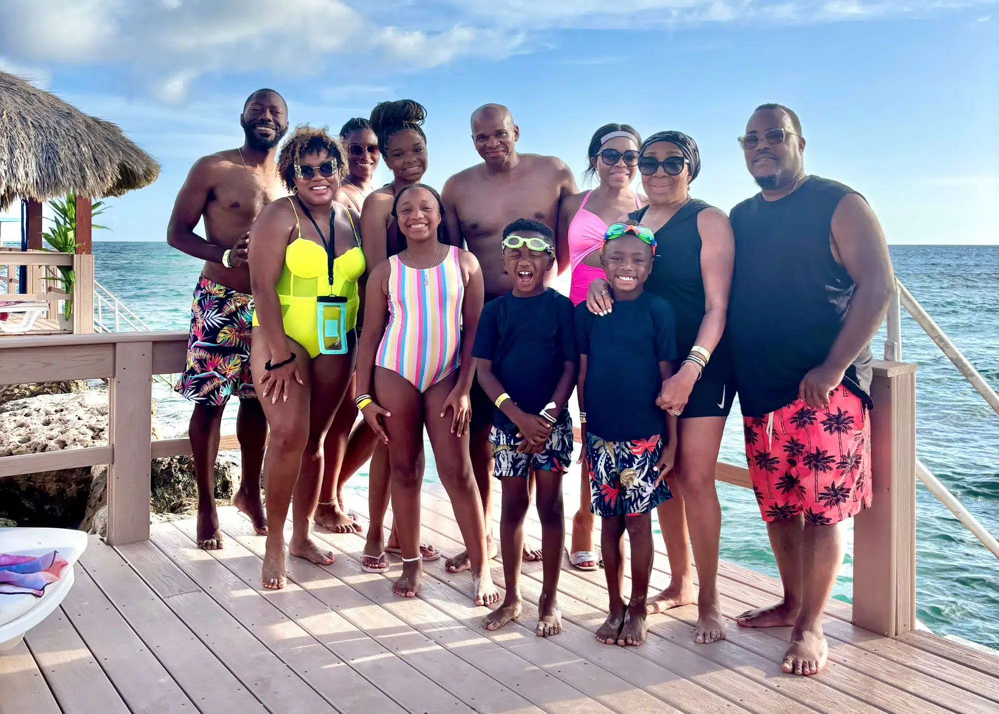 Marquita and her family pose for a group photo on a wooden deck overlooking the ocean at Flamingo Beach in Aruba. Everyone is dressed in swimwear, smiling and enjoying the tropical getaway together, with the clear blue sea and thatched beach hut in the background.