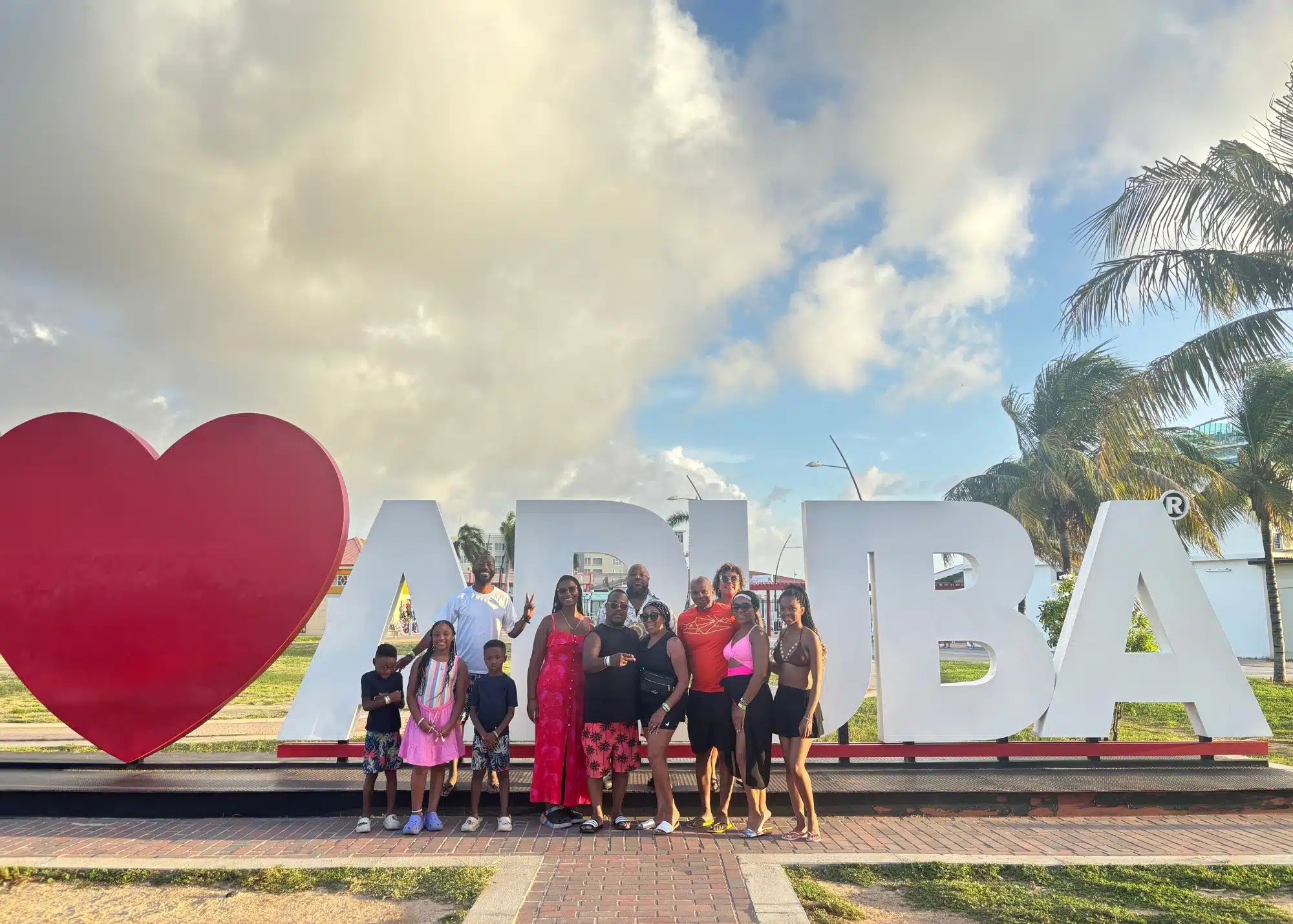 Marquita and her family posing in front of the large โI โค๏ธ Arubaโ sign under a partly cloudy sky, with palm trees and bright afternoon light in the background.
