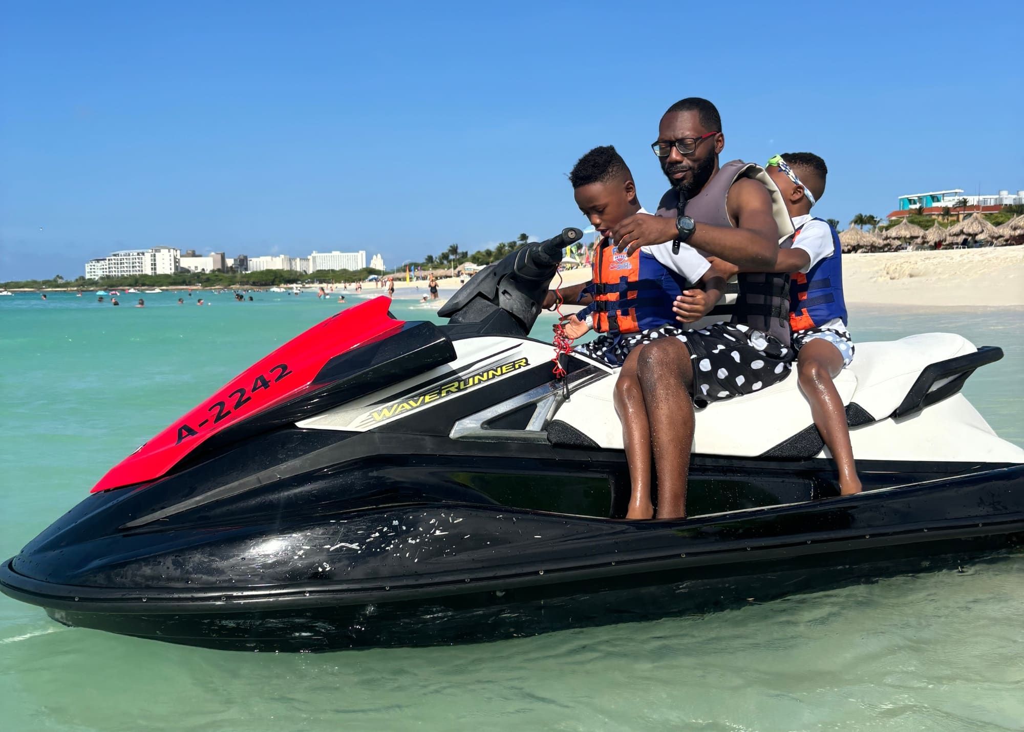 Marquita’s husband and their two sons riding a jet ski in shallow turquoise water, all wearing life vests. A lively beach scene and resort buildings in the background show the family-friendly fun at one of the best beaches in Aruba.