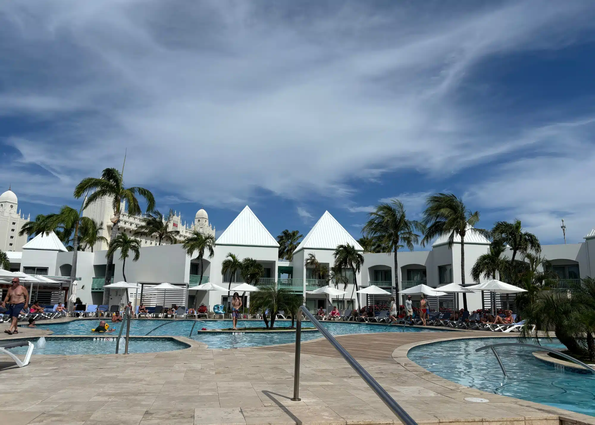 Wide view of the pool area at Courtyard Marriott in Aruba, with white modern buildings, palm trees, and people lounging under umbrellas.