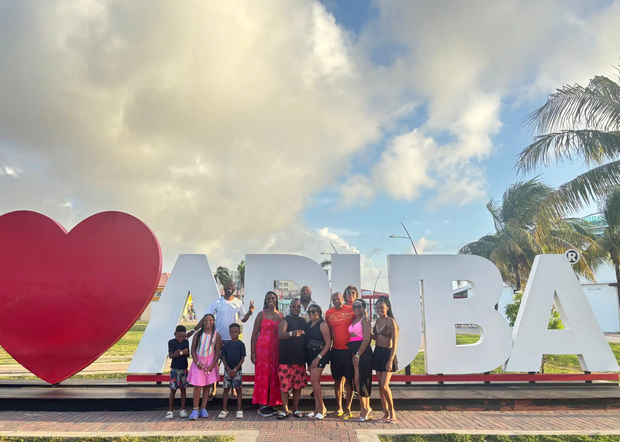Marquita and her family pose in front of the iconic “I ❤️ Aruba” sign under a partly cloudy sky, surrounded by palm trees and warm sunset light.