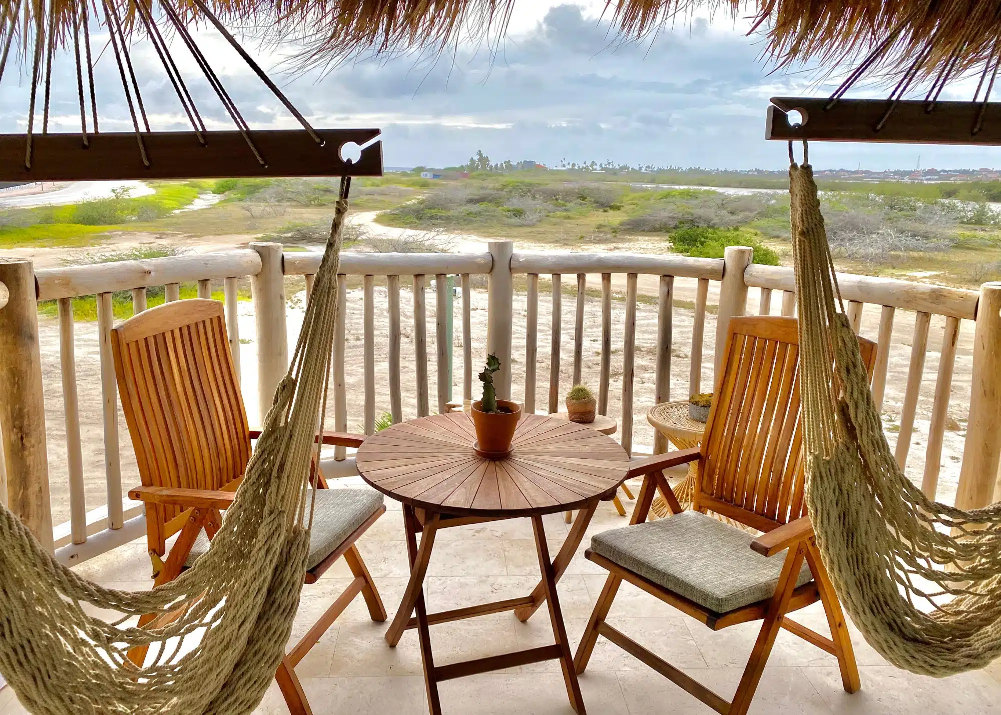 A cozy balcony setup at Boardwalk Boutique Hotel in Aruba features two wooden chairs, a round table with potted cactus plants, and two hanging rope hammocks overlooking a sandy, palm-dotted landscape.