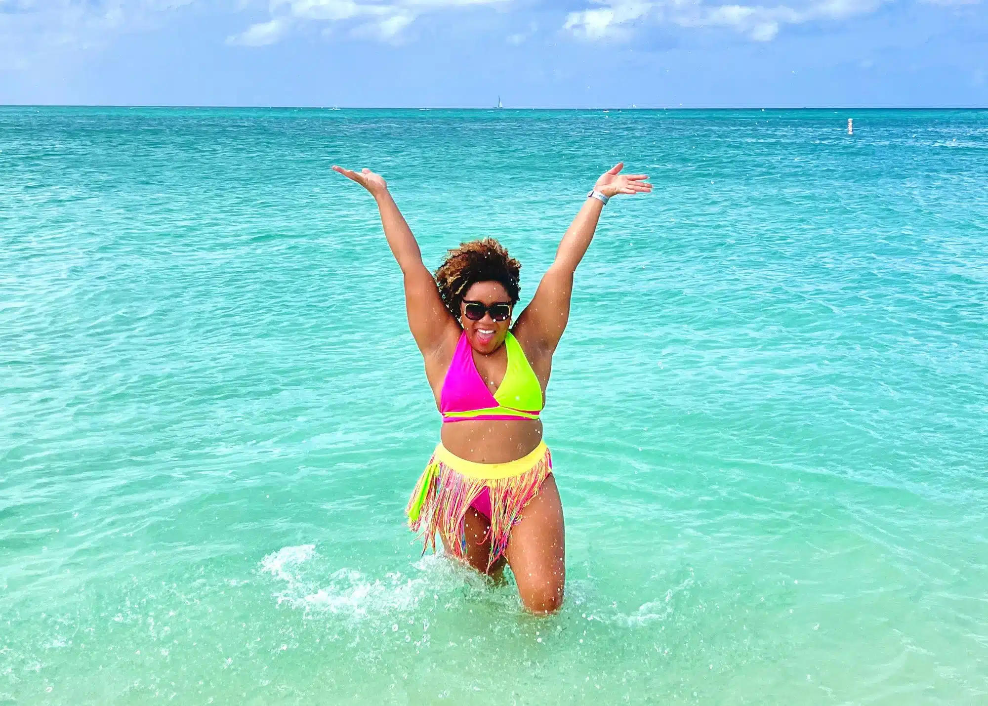 Marquita splashes in the clear turquoise water at Eagle Beach in Aruba, raising her arms with a big smile while wearing a colorful bikini and fringe skirt.
