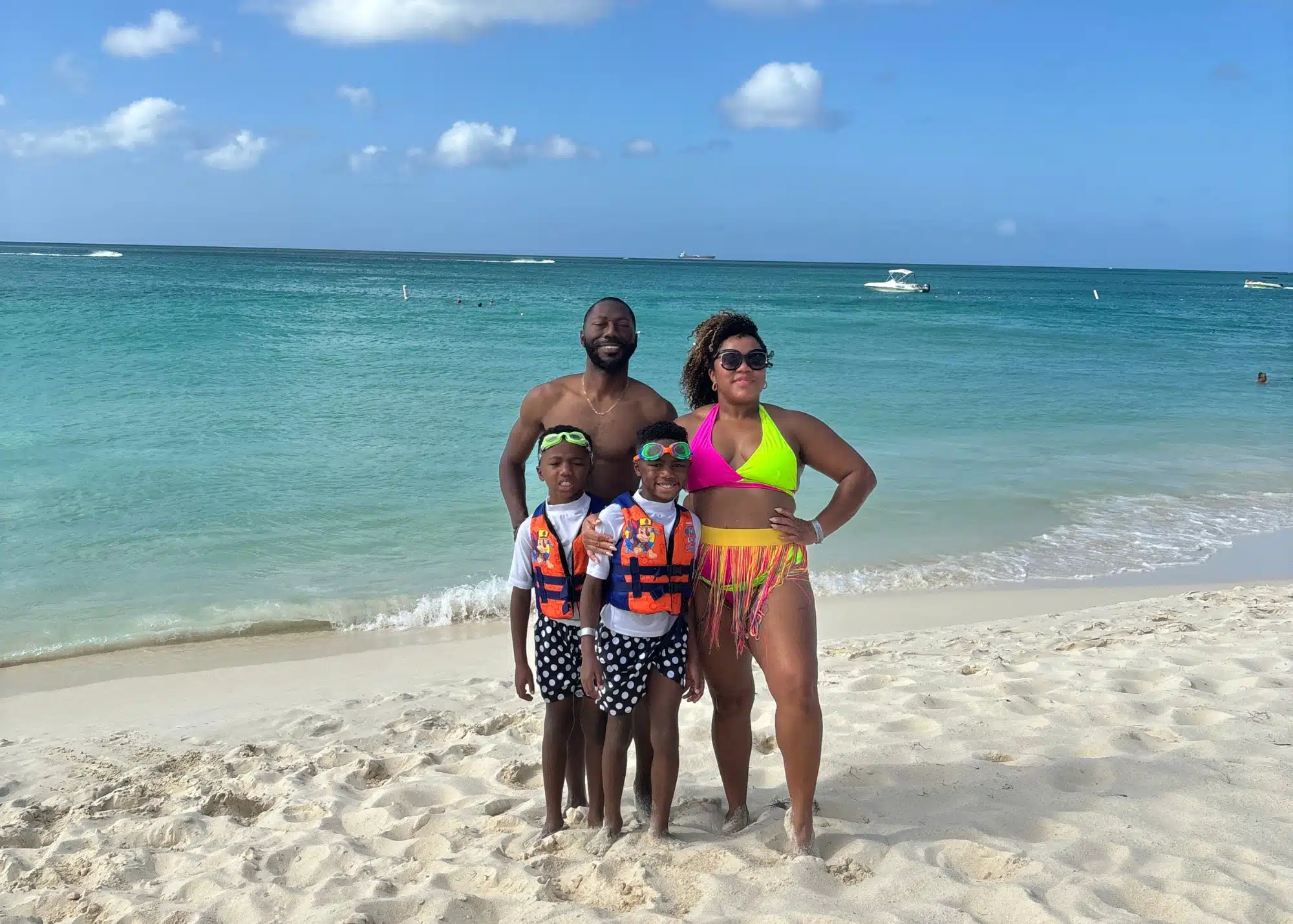 Marquita, her husband, and their two sons posing on the soft white sand of Eagle Beach in Aruba. With turquoise waves and boats in the distance, this family moment captures the relaxed vibe of one of the best beaches in Aruba.