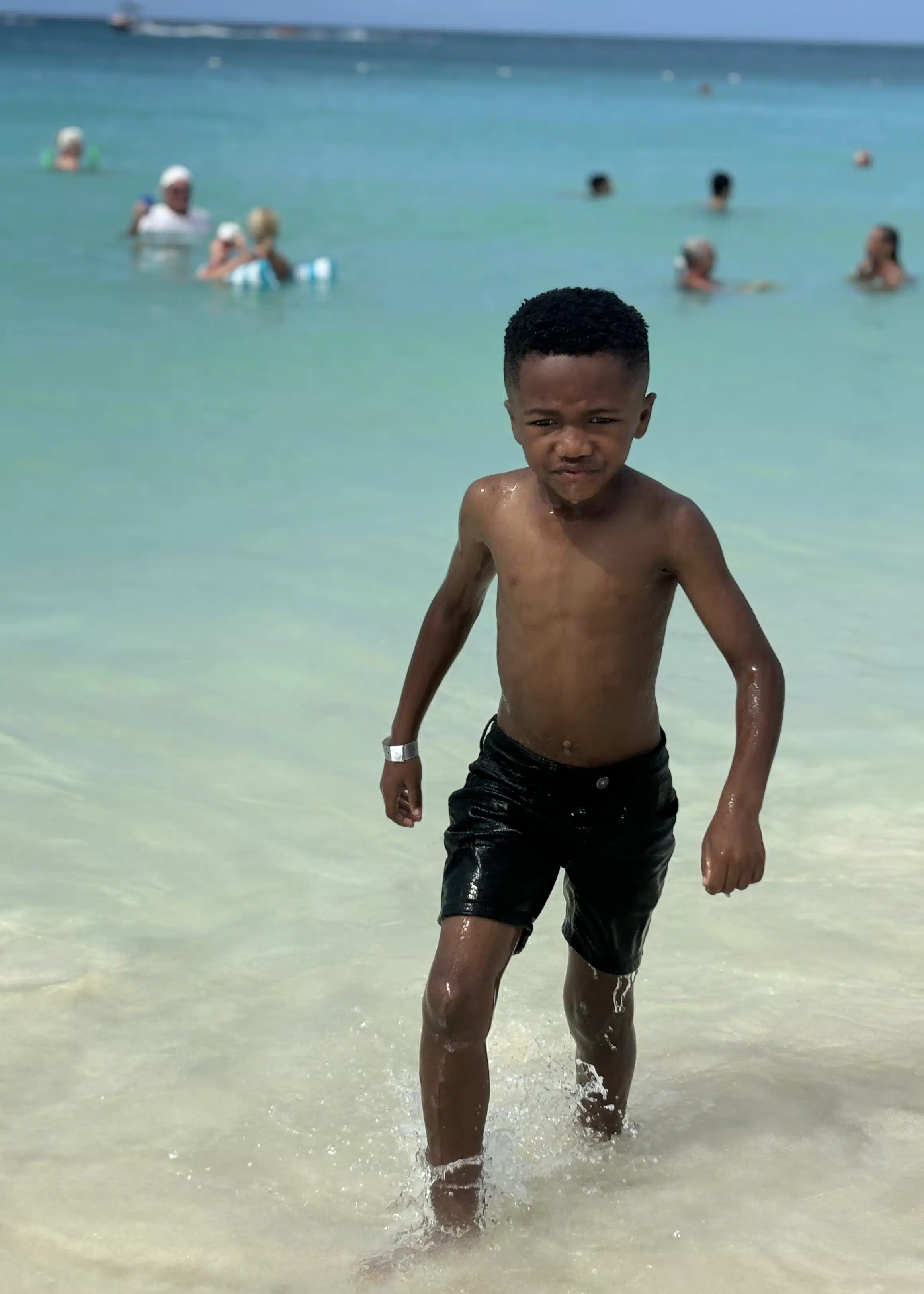 Marquita’s son walking out of the gentle surf at Eagle Beach in Aruba, with other beachgoers swimming in the turquoise water behind him.