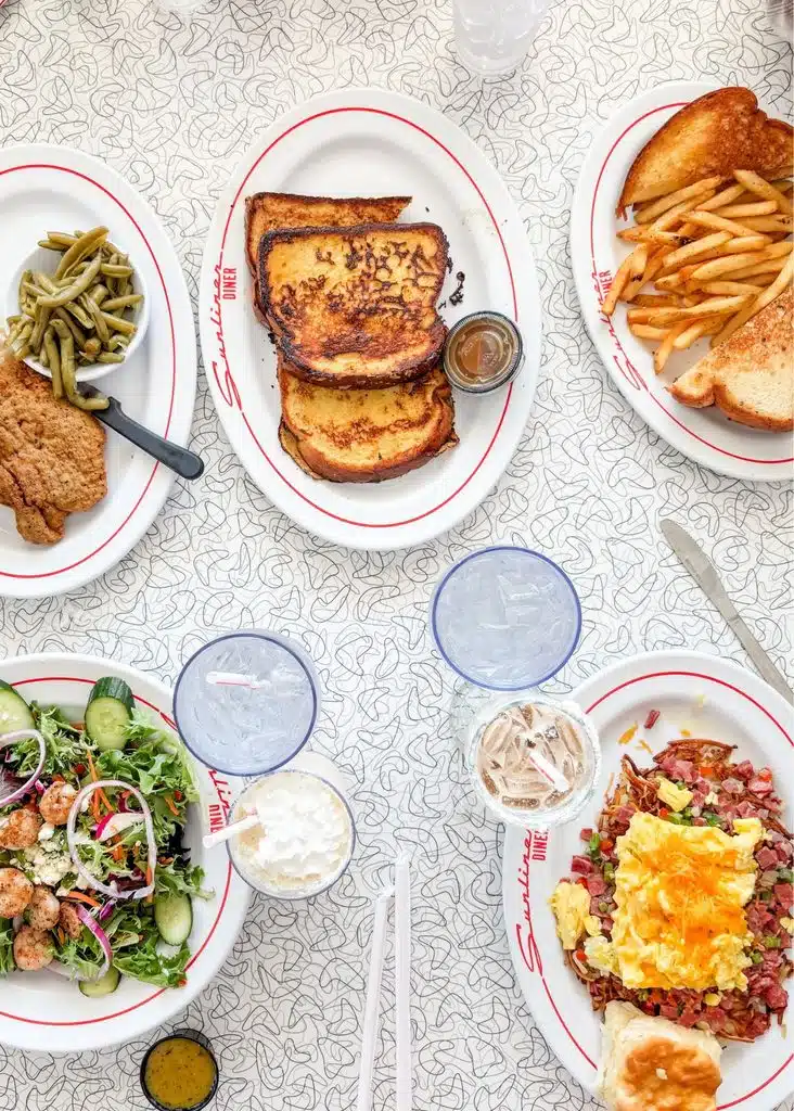 A colorful spread at Sunliner Diner includes French toast and fries alongside fried chicken with green beans and mashed potatoes. A shrimp salad and a hearty breakfast plate with eggs and corned beef hash round out the table.