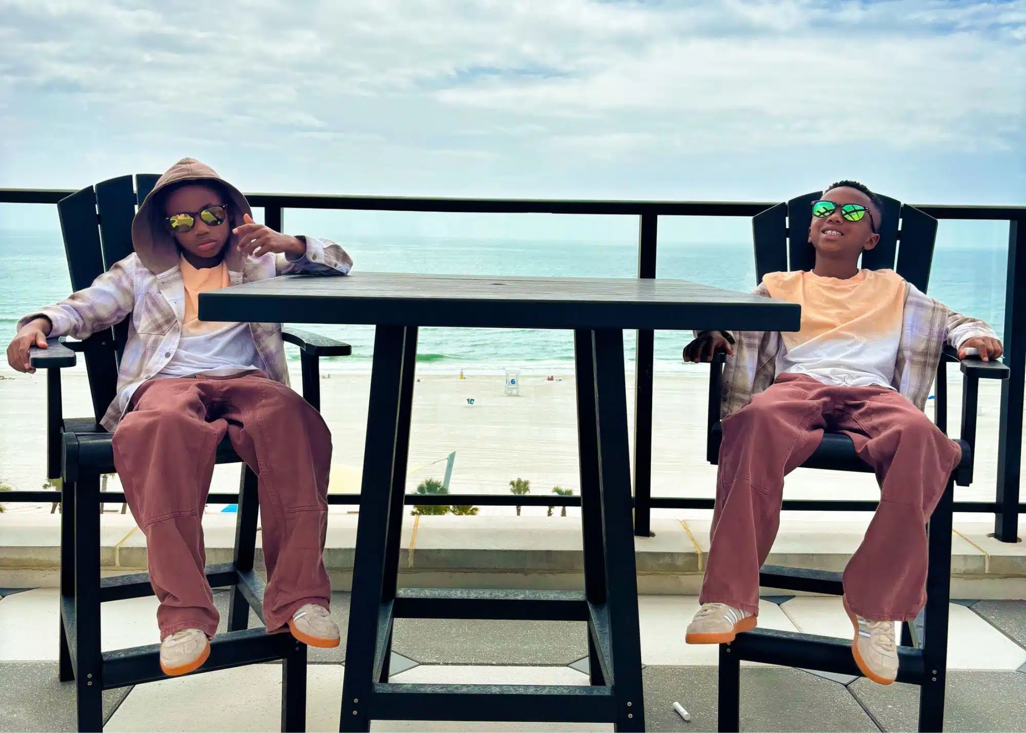 Marquita’s sons relax in tall patio chairs at their beachfront vacation rental in Gulf Shores Alabama. The balcony overlooks the white sand beach and Gulf beneath a partly cloudy sky.