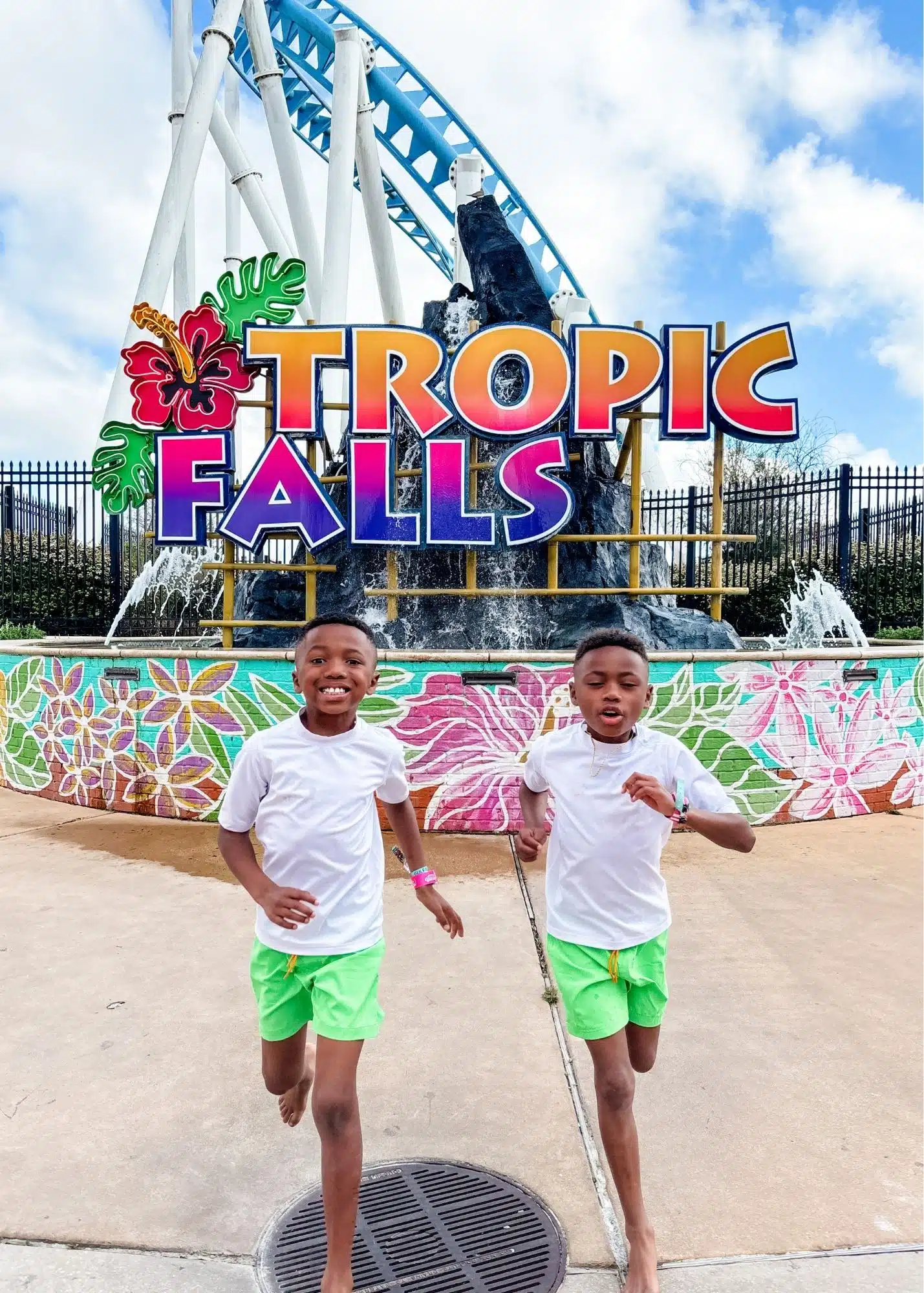 Marquita’s sons run barefoot in front of the colorful "Tropic Falls" sign at Tropic Falls at OWA. A blue roller coaster curves overhead behind them.