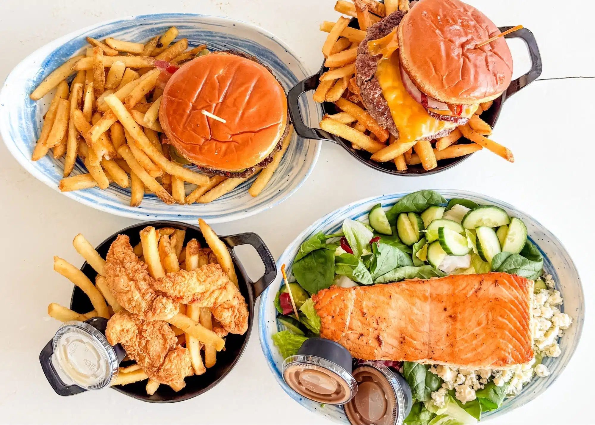 A spread of family friendly meals at The Hangout in Gulf Shores Alabama with cheeseburgers and fries. Fried chicken tenders and fries sit beside a grilled salmon salad topped with cucumbers and crumbled cheese.