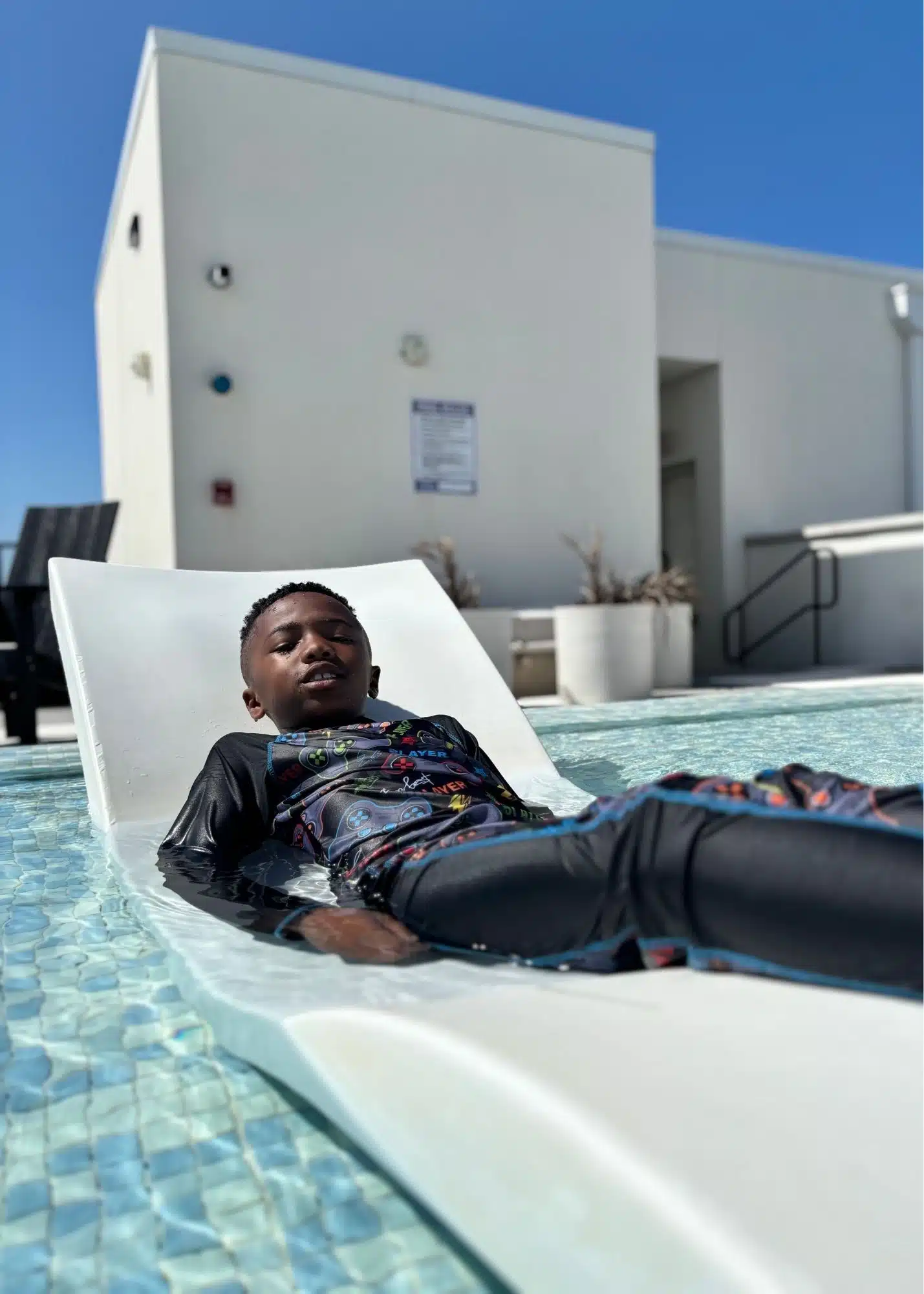 One of Marquita’s sons reclines on a submerged lounge chair in the rooftop pool at their beachfront vacation rental. The shallow water surrounds the chair beneath a bright blue sky.