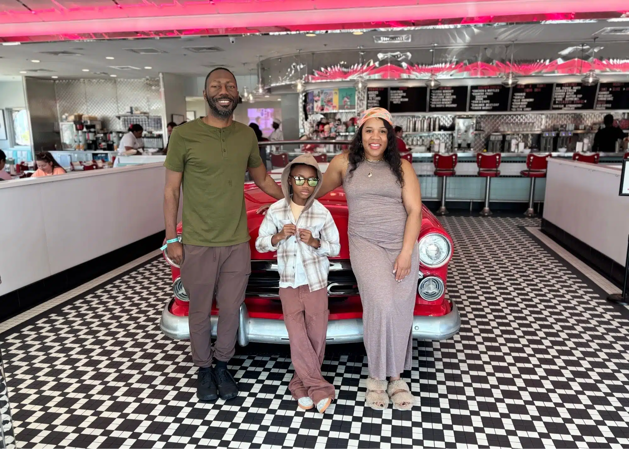 Marquita poses with her husband and one of her sons in front of a vintage red car inside the Sunliner Diner. The retro restaurant features a black and white checkered floor and classic diner counter.