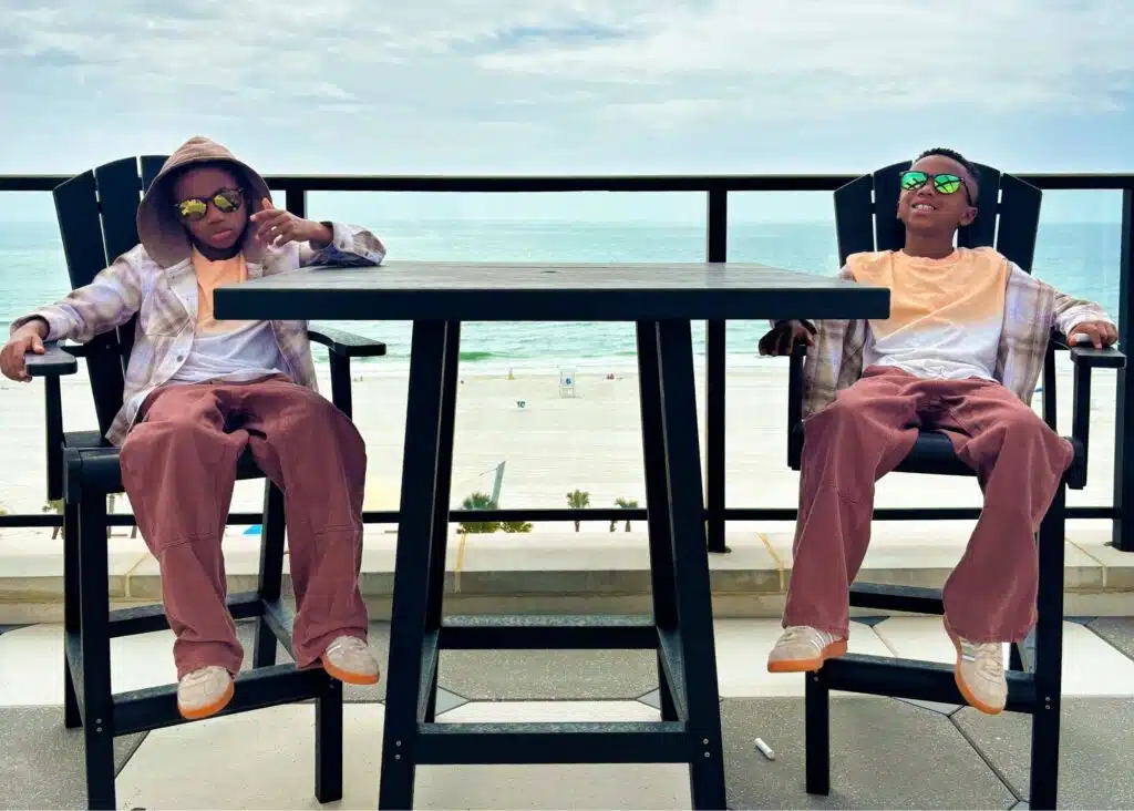 Marquita’s sons relax in tall patio chairs at their beachfront vacation rental in Gulf Shores Alabama. The balcony overlooks the white sand beach and Gulf beneath a partly cloudy sky.