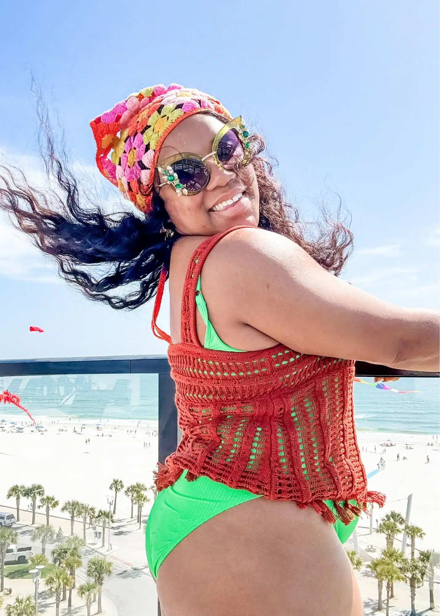 Marquita smiles on a balcony overlooking the white sand beach and Gulf in Gulf Shores Alabama. She wears a bright green swimsuit with a colorful crochet head wrap and cover up as the wind blows through her hair.