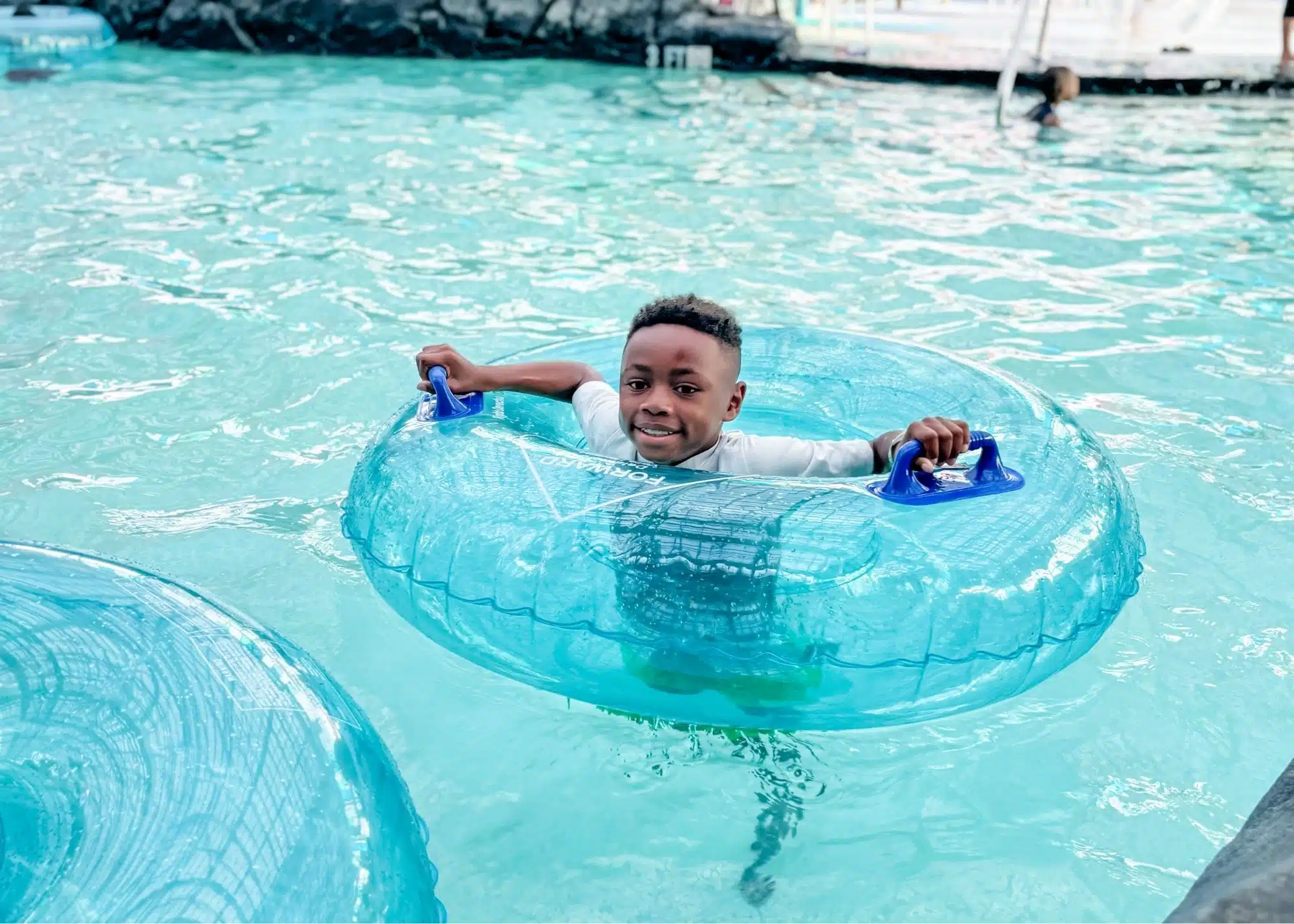 One of Marquita’s sons floats in a blue inner tube through the turquoise water at Tropic Falls. Colorful umbrellas and tropical themed surroundings fill the water park behind him.