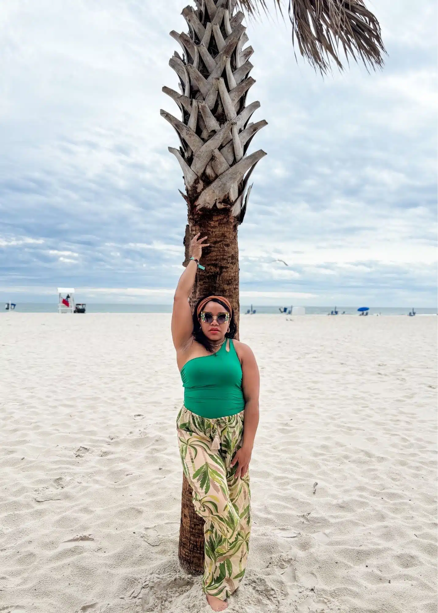 Marquita poses against a palm tree on the white sand beach in Gulf Shores Alabama. She wears a green top and tropical print pants with the Gulf of Mexico stretching behind her.