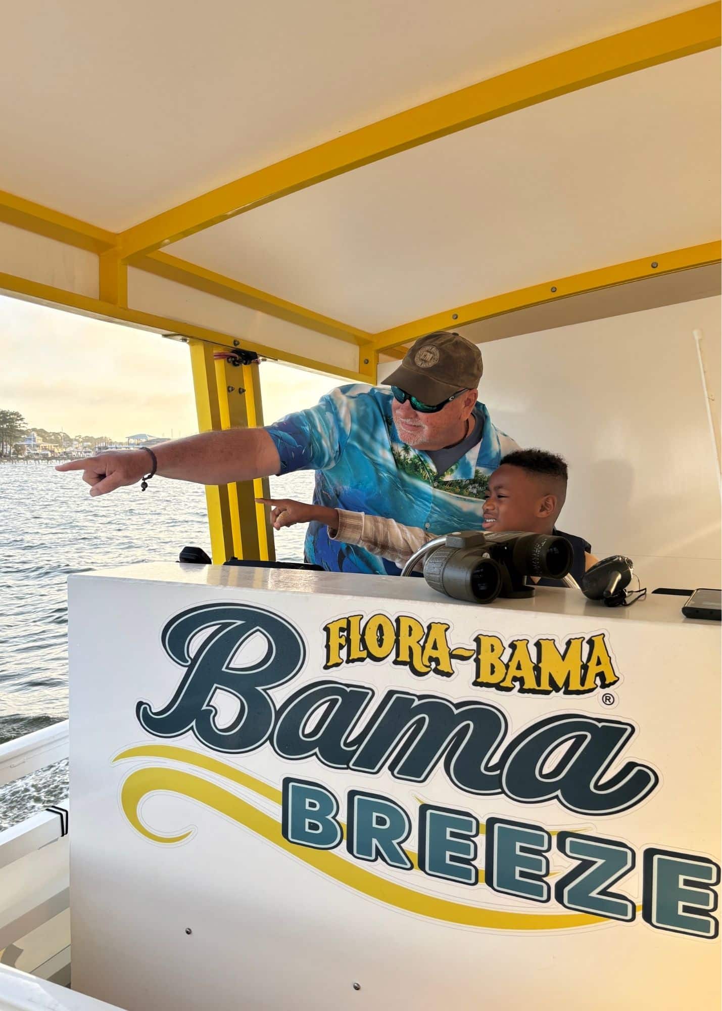 One of Marquita’s sons points across the water with a crew member aboard the Bama Breeze Dolphin Cruise from Flora Bama Marina. Binoculars rest nearby as they look for dolphins.