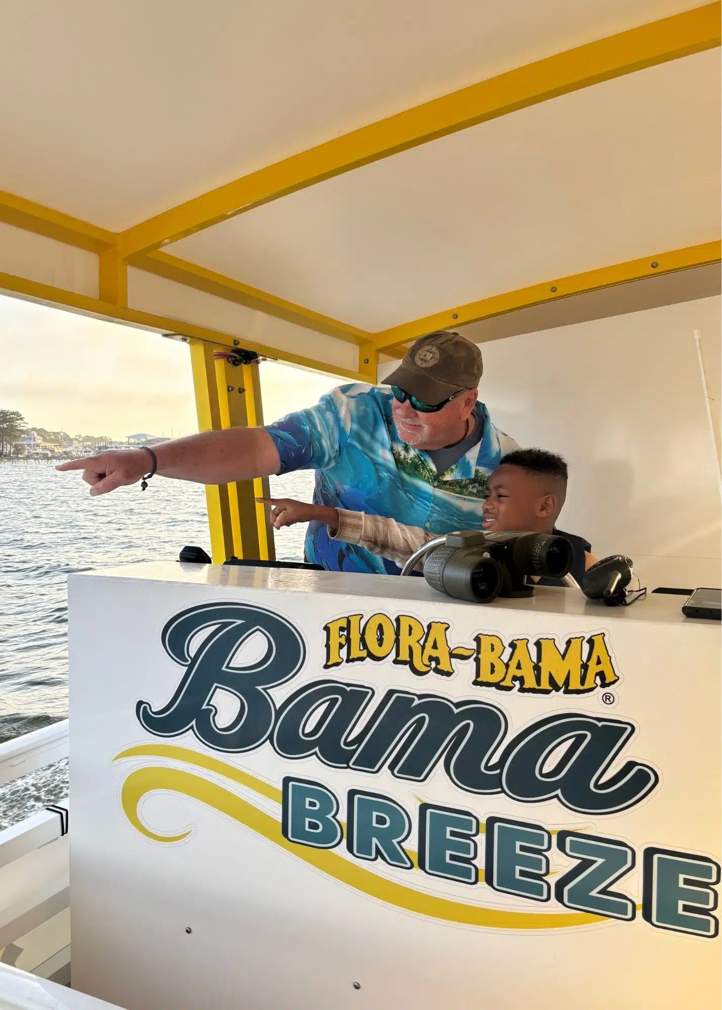 One of Marquita’s sons points across the water with a crew member aboard the Bama Breeze Dolphin Cruise from Flora Bama Marina. Binoculars rest nearby as they look for dolphins.