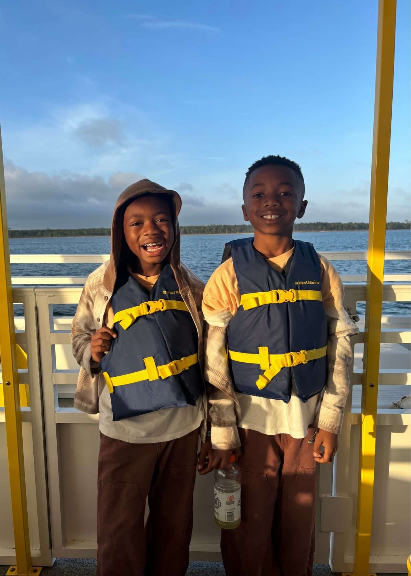 Marquita’s sons smile together in life jackets aboard the Bama Breeze Dolphin Cruise from Flora Bama Marina. Calm blue water stretches behind them in the evening light.