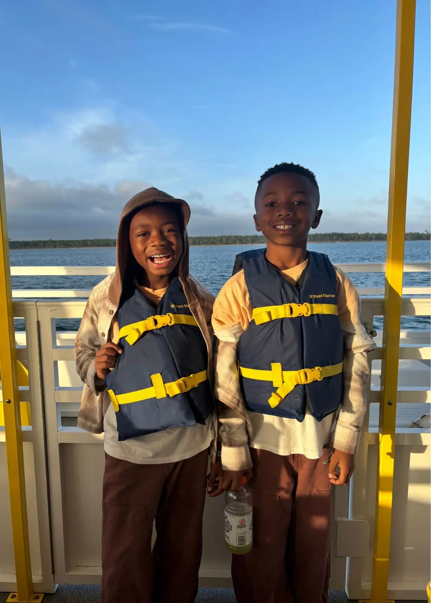 Marquita’s sons smile together in life jackets aboard the Bama Breeze Dolphin Cruise from Flora Bama Marina. Calm blue water stretches behind them in the evening light.