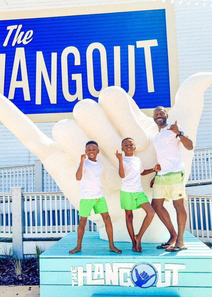 Marquita’s sons pose with Marquita’s husband in front of a giant shaka hand sculpture at The Hangout in Gulf Shores Alabama. All three smile and make shaka hand signs beneath the large "The Hangout" sign.