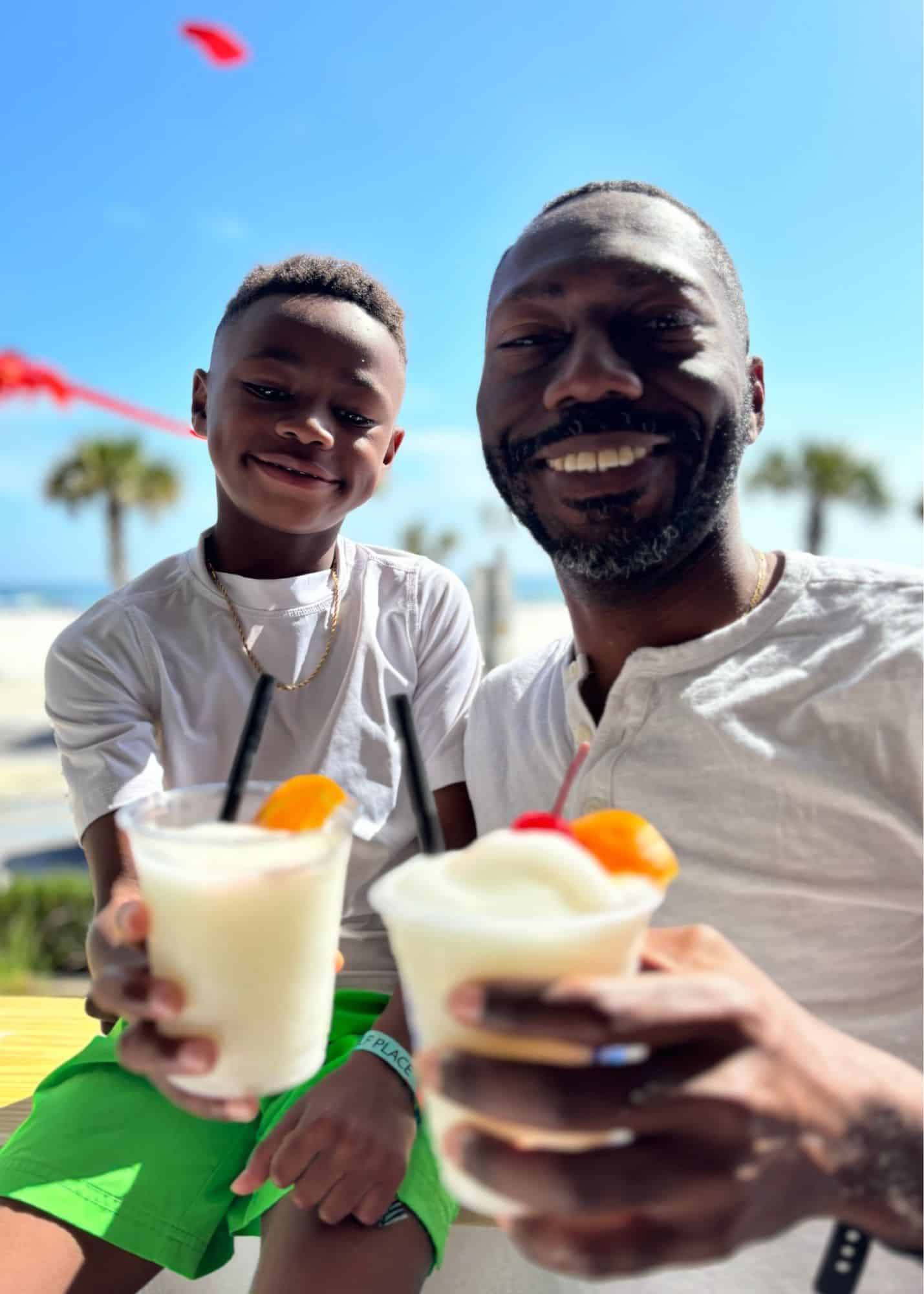 One of Marquita’s sons and Marquita’s husband smile together while enjoying frozen drinks at The Hangout in Gulf Shores Alabama. Palm trees and the beach are visible behind them.