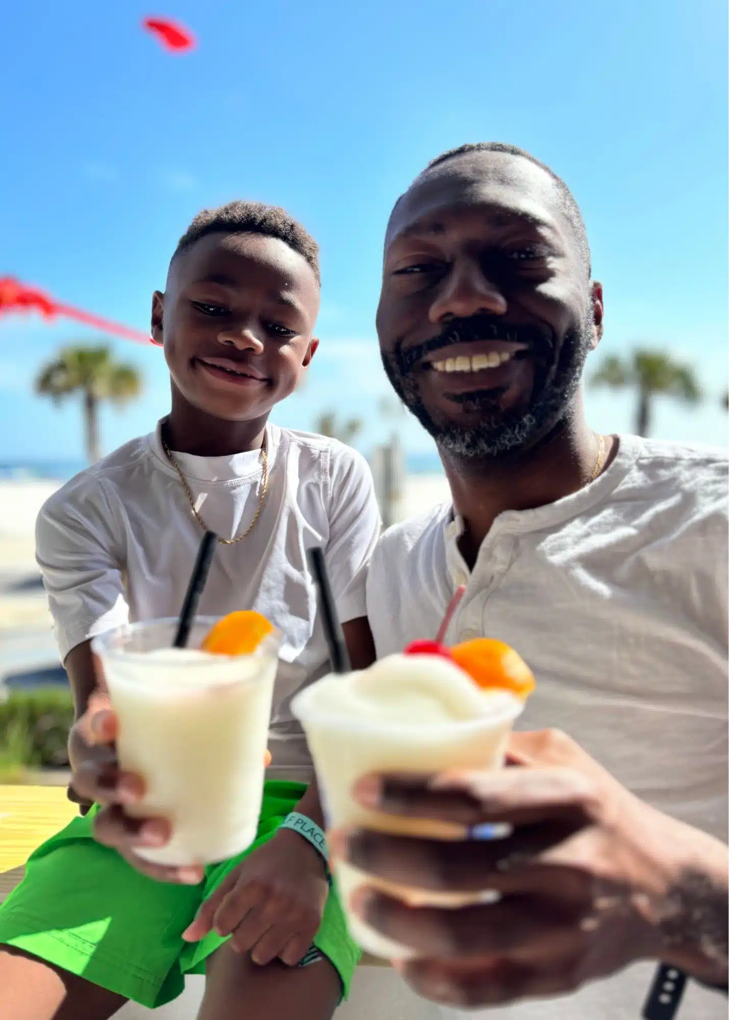 One of Marquita’s sons and Marquita’s husband smile together while enjoying frozen drinks at The Hangout in Gulf Shores Alabama. Palm trees and the beach are visible behind them.