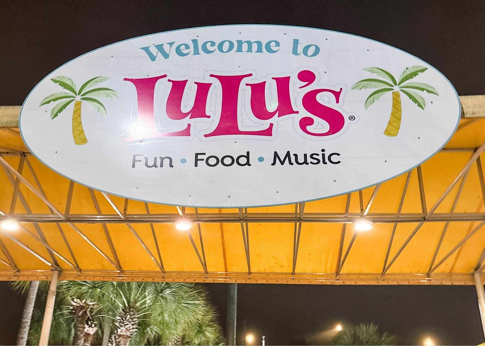 The entrance to LuLu’s in Gulf Shores Alabama at night with palm trees and outdoor seating visible beyond the entryway. A large sign overhead reads "Welcome to LuLu’s Fun Food Music."