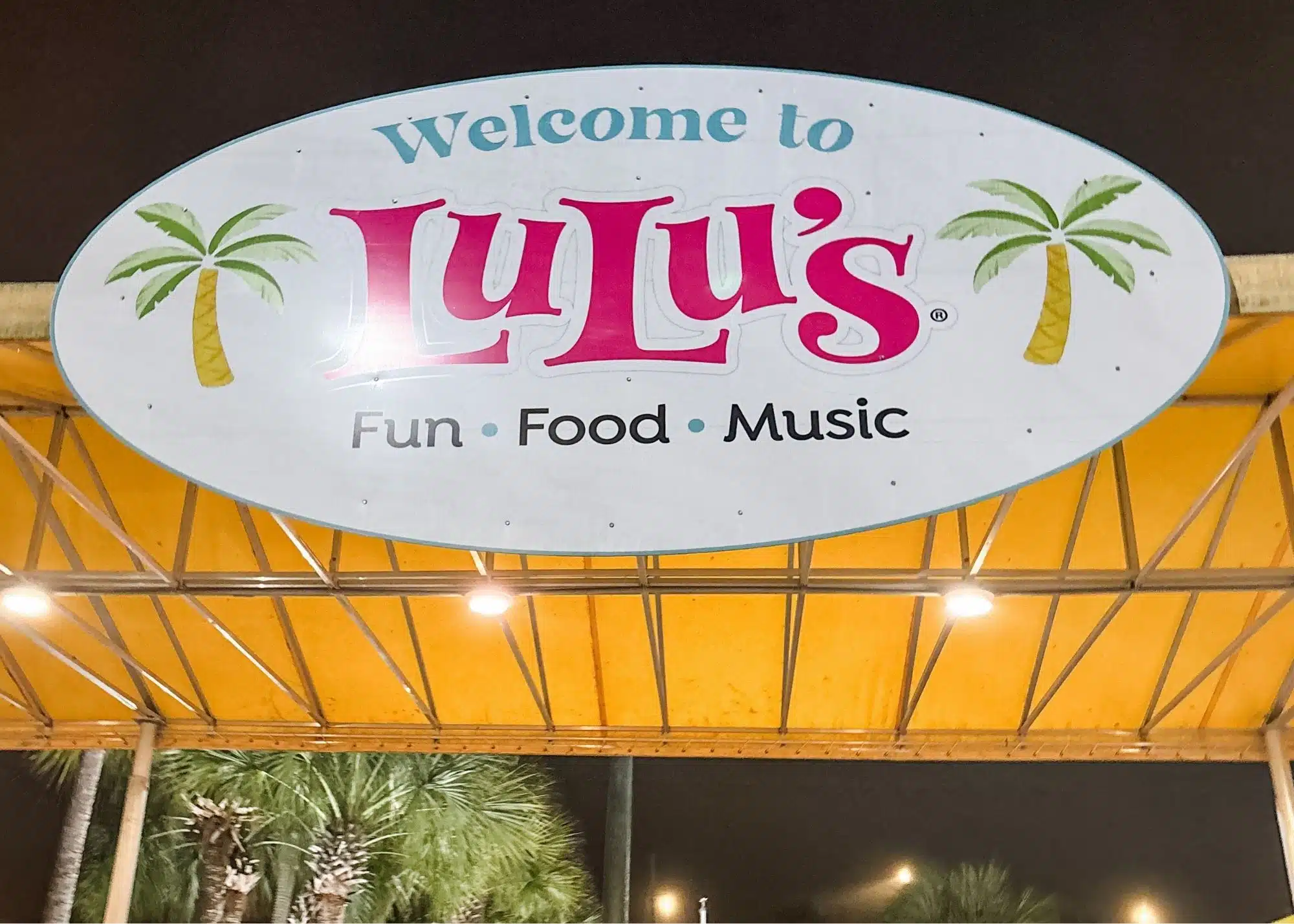 The entrance to LuLu’s in Gulf Shores Alabama at night with palm trees and outdoor seating visible beyond the entryway. A large sign overhead reads "Welcome to LuLu’s Fun Food Music."