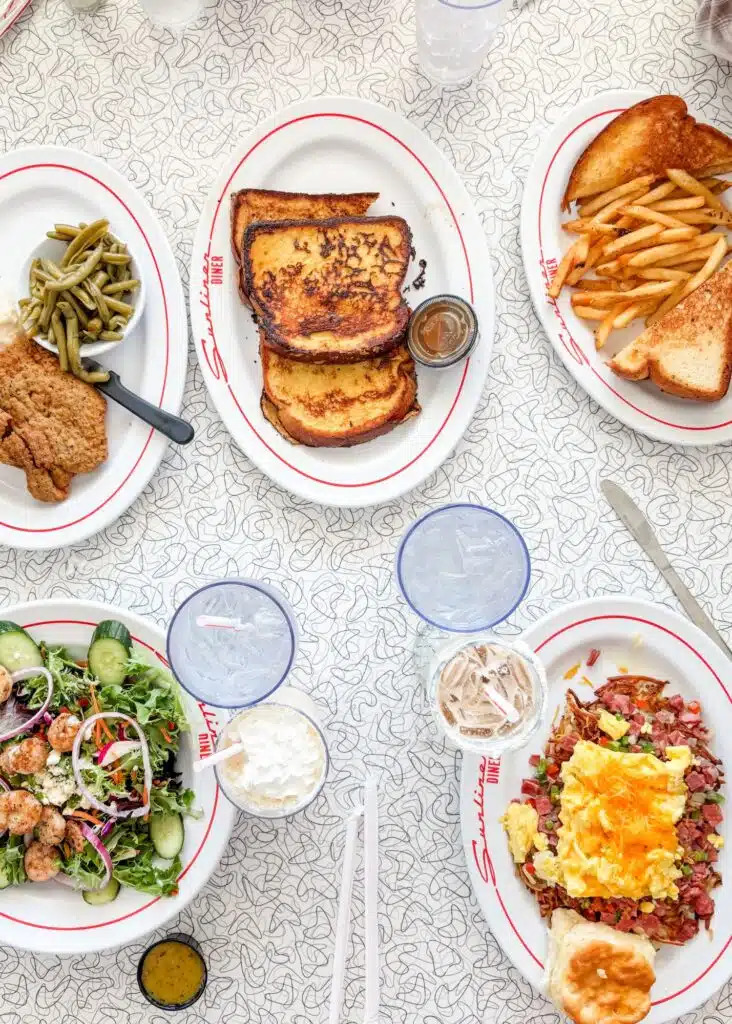 A colorful spread at Sunliner Diner includes French toast and fries alongside fried chicken with green beans and mashed potatoes. A shrimp salad and a hearty breakfast plate with eggs and corned beef hash round out the table.