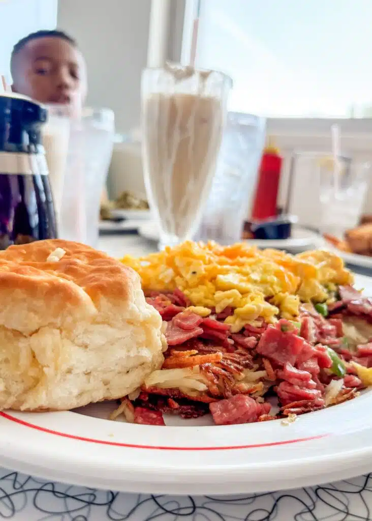 A hearty breakfast at Sunliner Diner features scrambled eggs with melted cheese over corned beef hash alongside a large fluffy biscuit. A tall creamy milkshake is visible on the table in the background