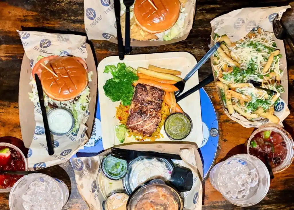 An overhead spread of food from The Gulf with burgers and grilled fish served with vegetables and rice. Parmesan topped fries and several dipping sauces fill the table alongside the entrees.
