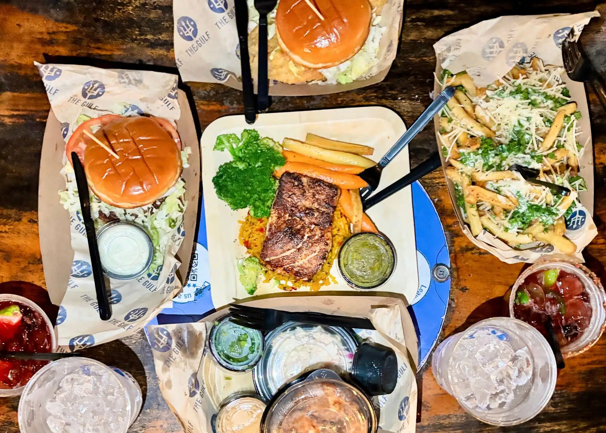 An overhead spread of food from The Gulf with burgers and grilled fish served with vegetables and rice. Parmesan topped fries and several dipping sauces fill the table alongside the entrees.