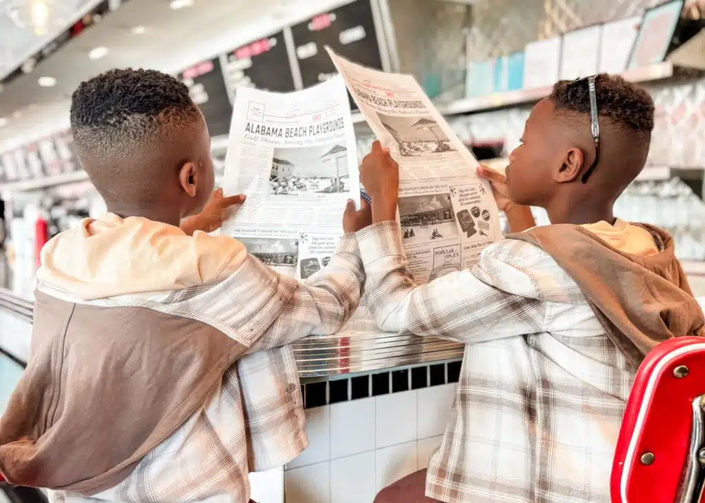 Marquita’s sons sit on red stools at the counter inside Sunliner Diner while reading menus designed like vintage newspapers. The retro diner has chrome details and classic black and white tile.