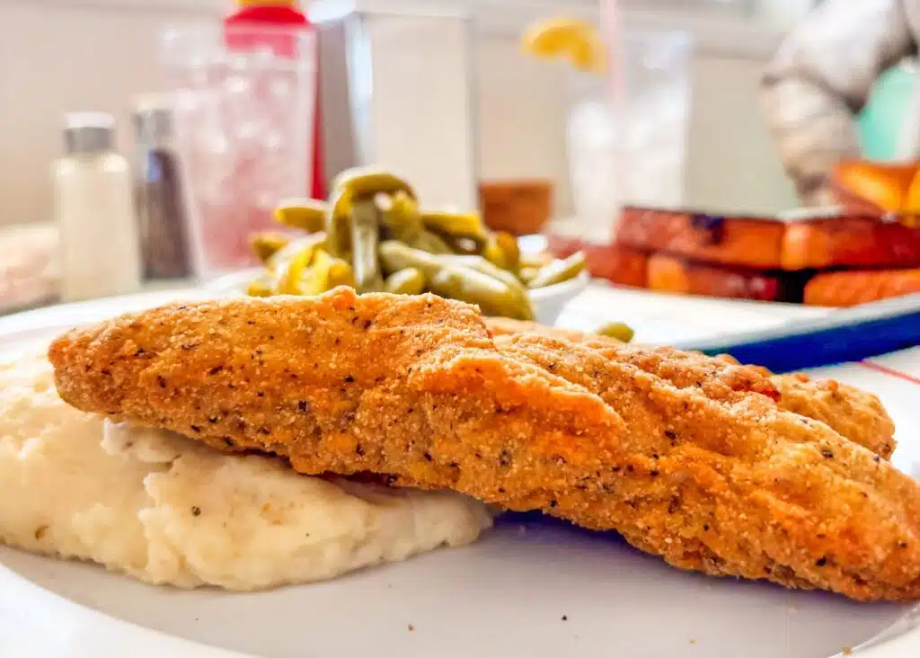 A crispy fried fish fillet from Sunliner Diner rests over creamy mashed potatoes with green beans served behind it. The golden breading and generous portion fill most of the plate.