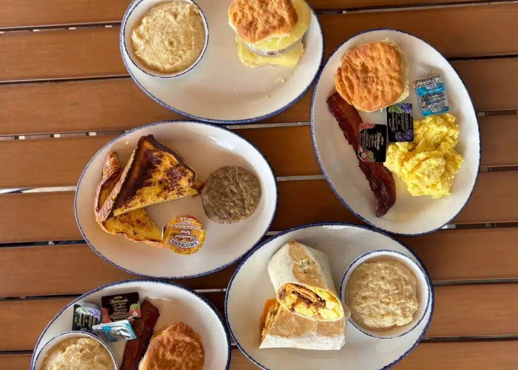 A breakfast spread at CoastAL Orange Beach includes biscuits with grits and bacon alongside scrambled eggs. French toast with sausage and a breakfast burrito with grits complete the table.