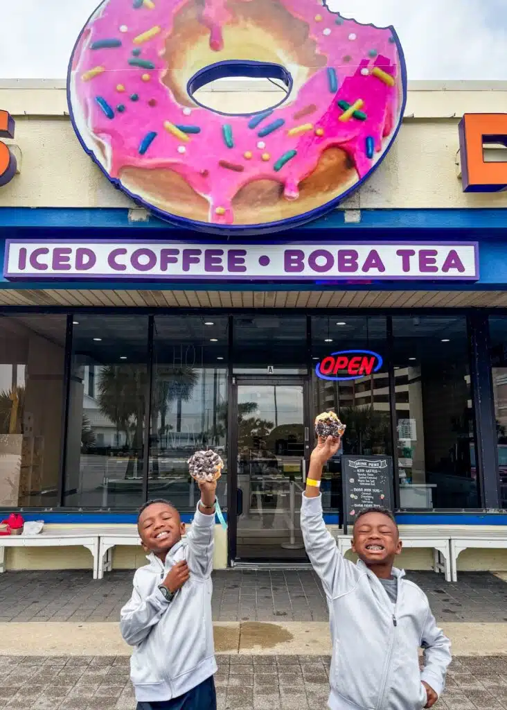 Marquita’s sons smile outside Daisy’s Donuts while holding up their chocolate covered donuts beneath a giant pink sprinkled donut sign. The storefront sign reads "ICED COFFEE. BOBA TEA."